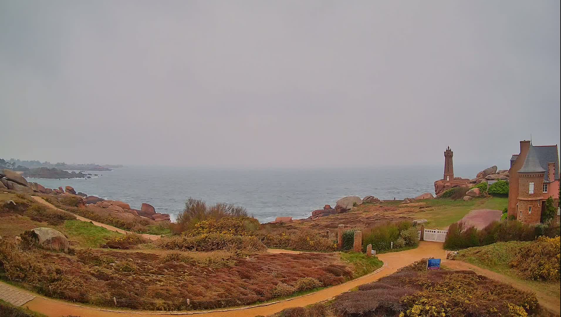A winding path leads to a stone house and a lighthouse overlooking a calm, gray sea under an overcast sky.