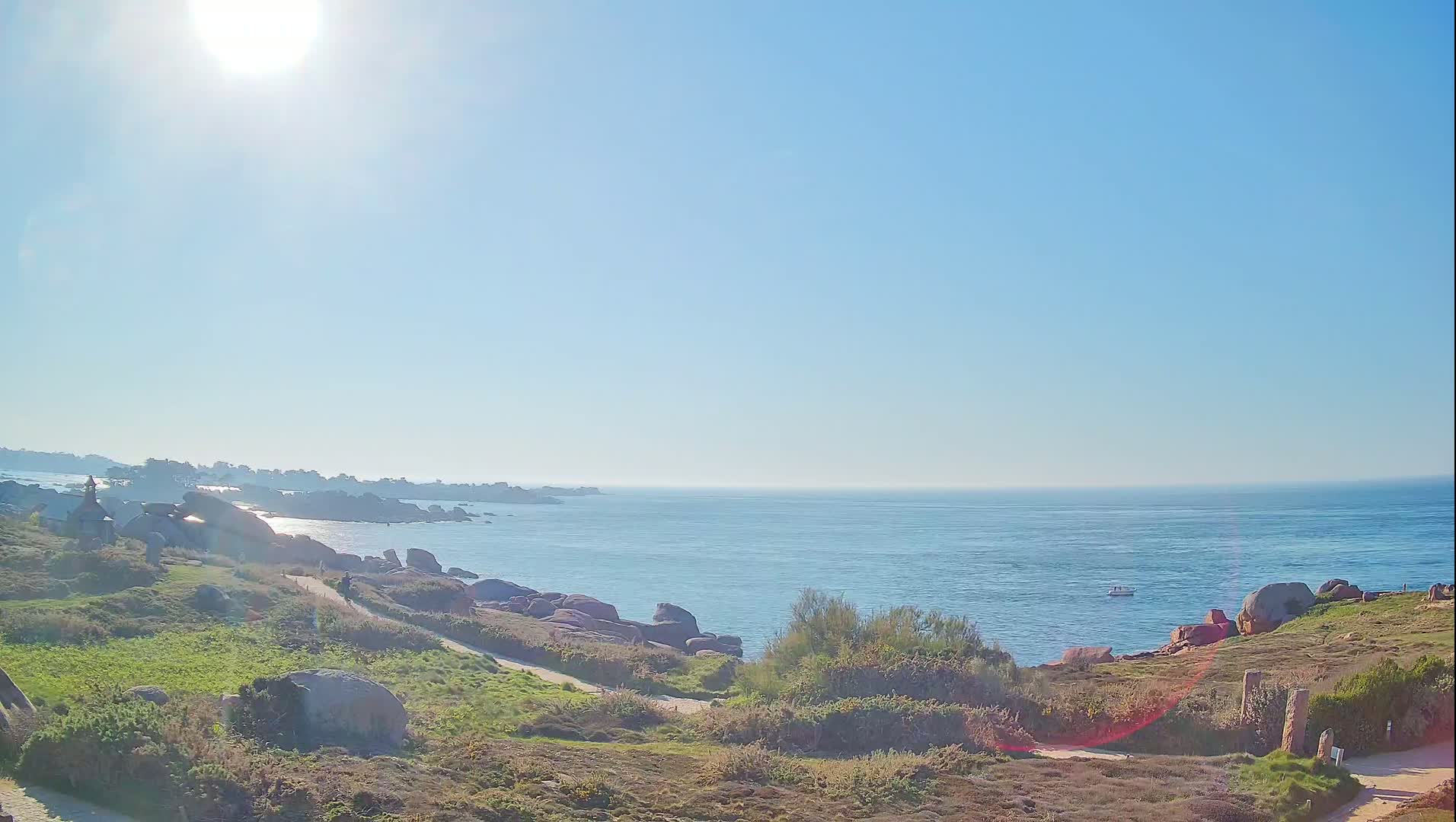 A sunny day reveals a coastal scene with a calm ocean, rocky shoreline, grassy areas, and a person walking along a path.