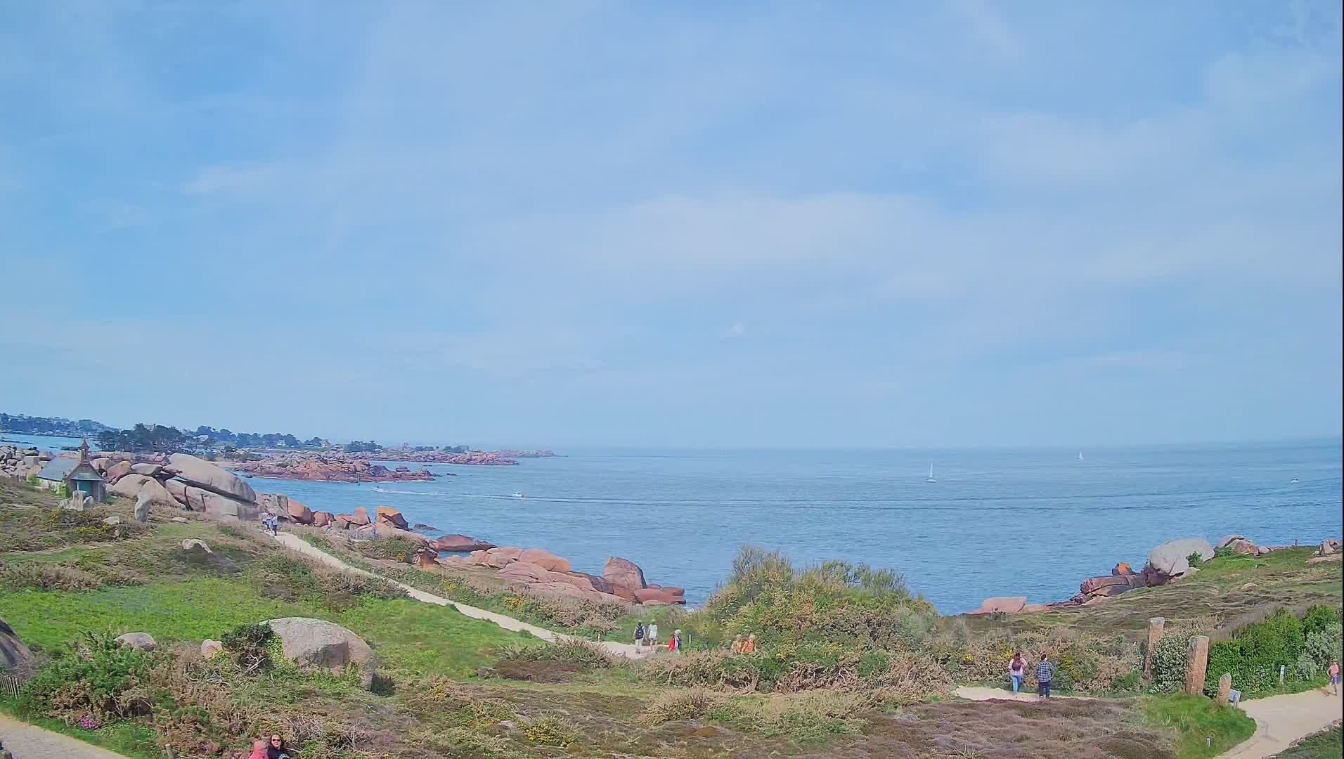 A coastal scene under a mostly clear sky shows a rocky shoreline with a few people walking along a path overlooking a calm ocean with a few boats in the distance.