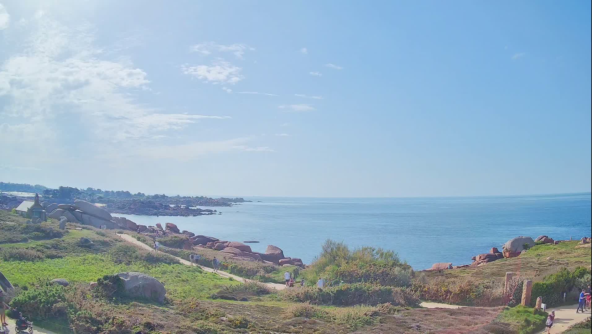 A coastal path winds through grassy terrain dotted with large pink rocks, leading to a calm ocean under a mostly sunny sky with scattered clouds.