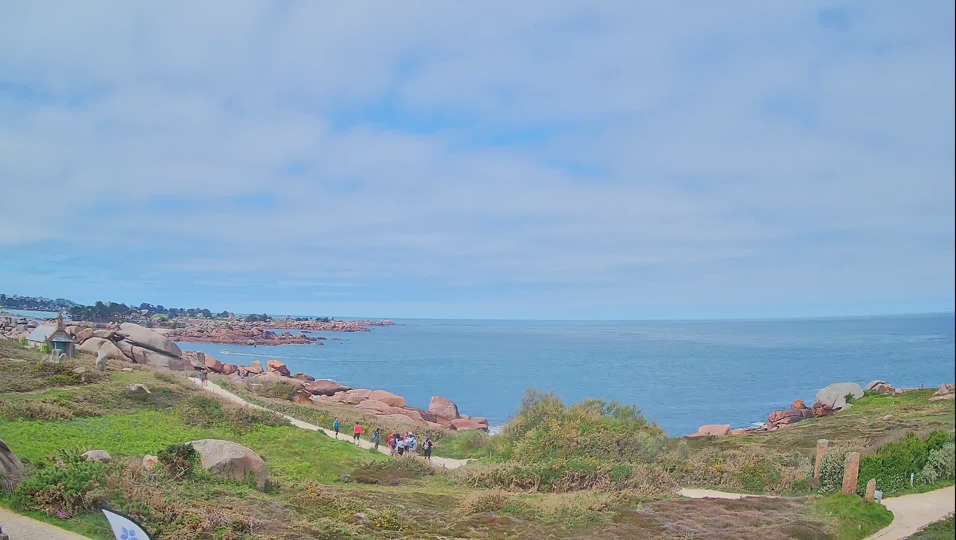 A coastal path winds through grassy terrain dotted with pink granite boulders, overlooking a calm sea under a partly cloudy sky.