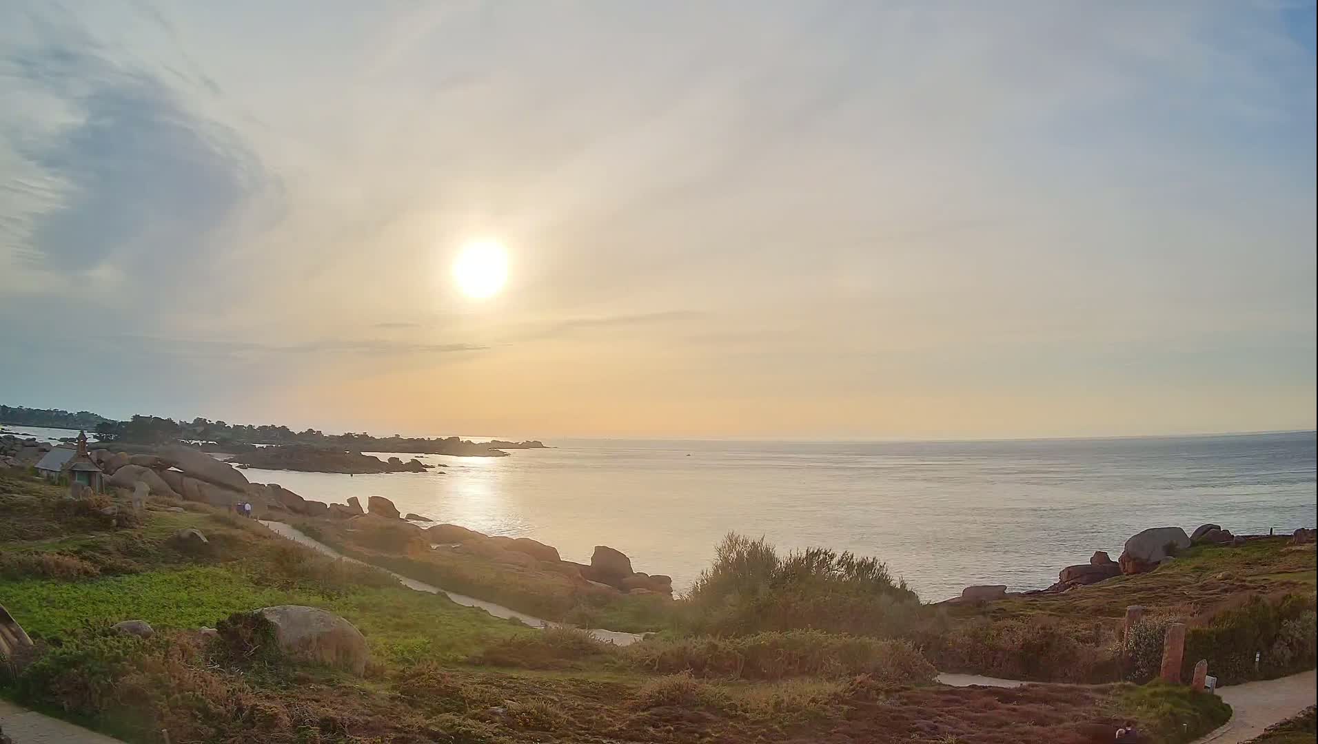 A calm ocean at sunset is viewed from a grassy hillside dotted with large rocks and a path, under a partly cloudy sky.