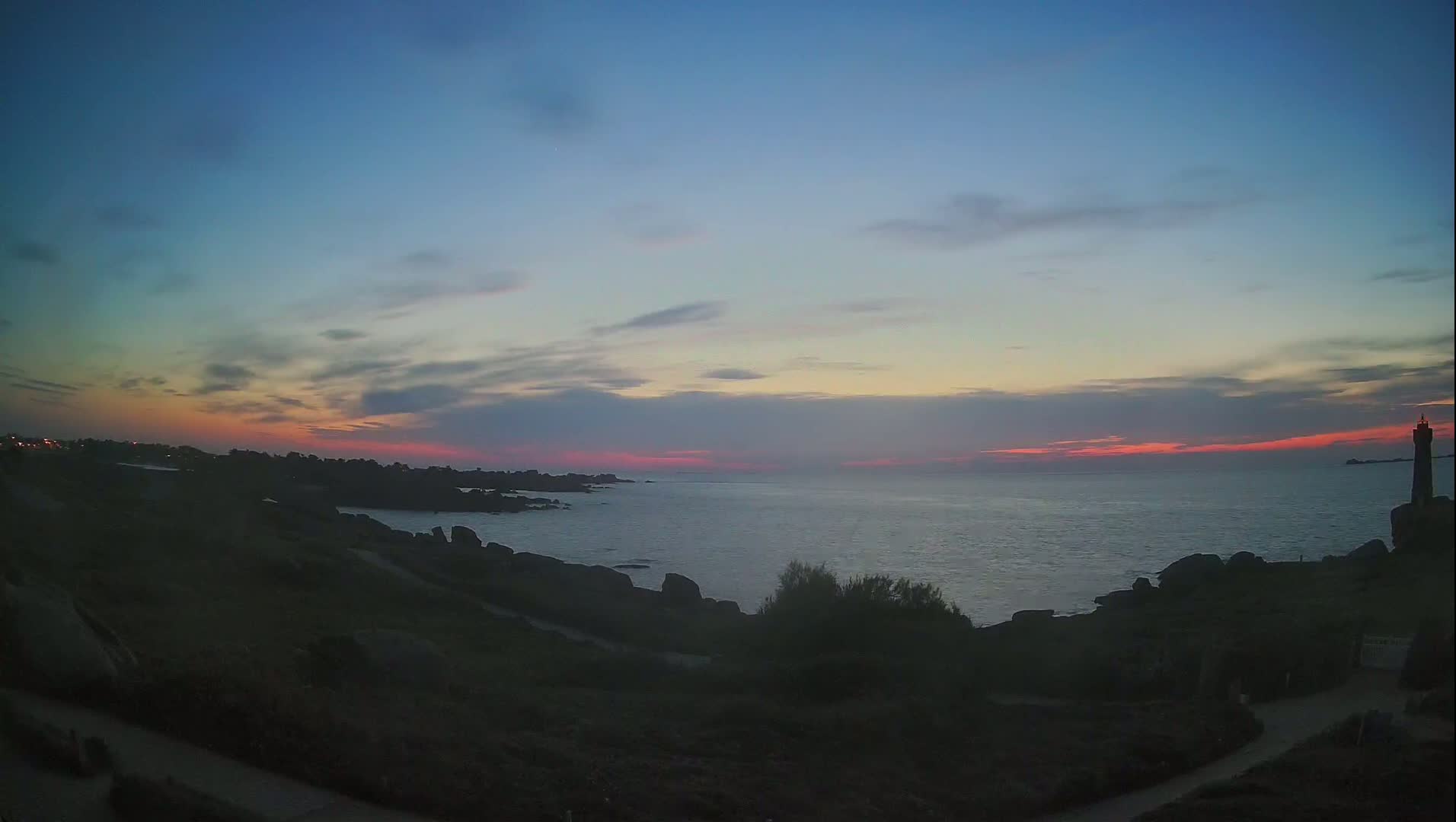 A rocky coastline at sunset, under a mostly clear sky with pink and purple hues on the horizon, features a lone lighthouse in the distance.