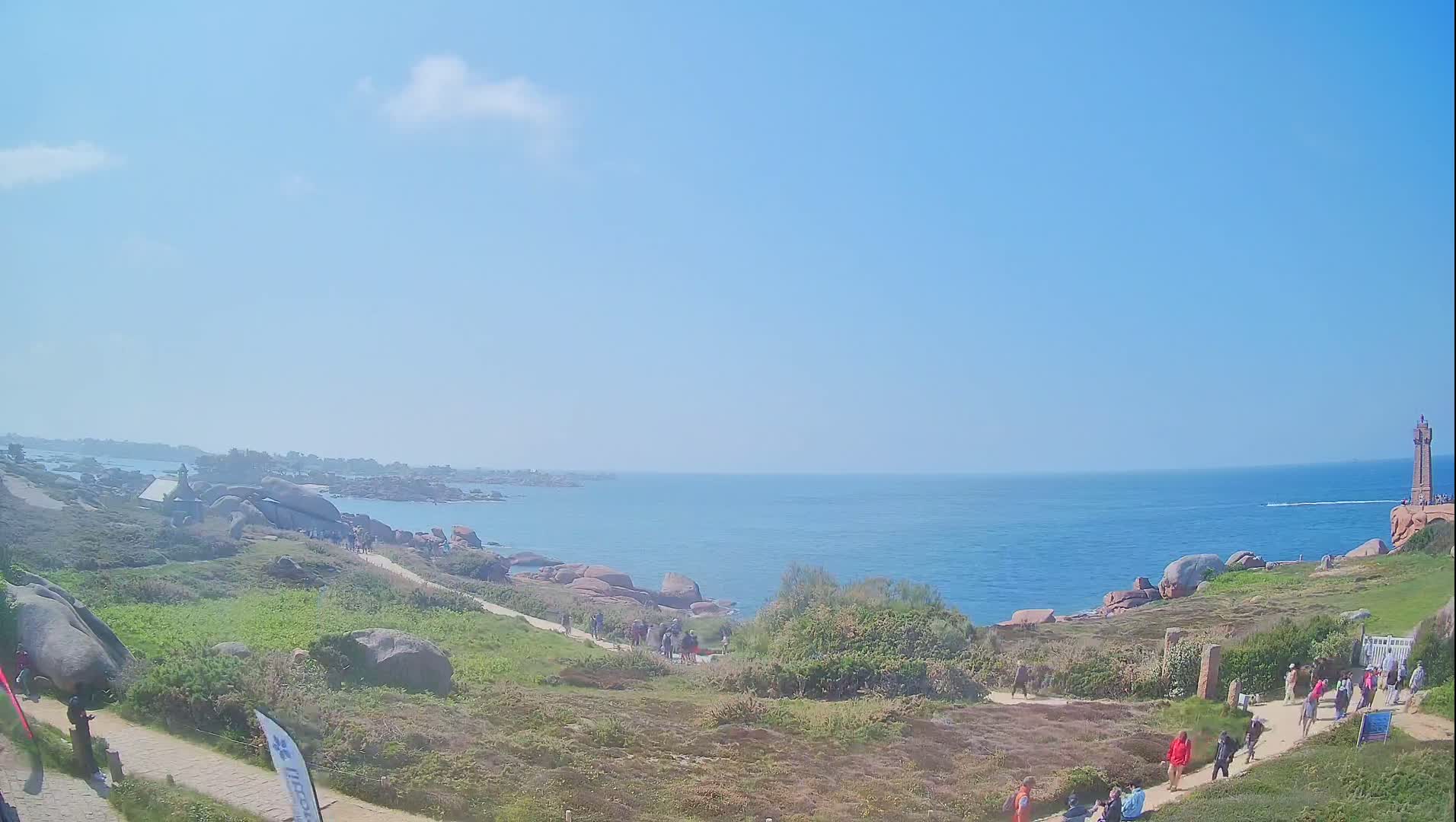 A coastal path winds through a rocky landscape overlooking a calm blue sea under a mostly clear sky, with people walking along the path toward a distant lighthouse.