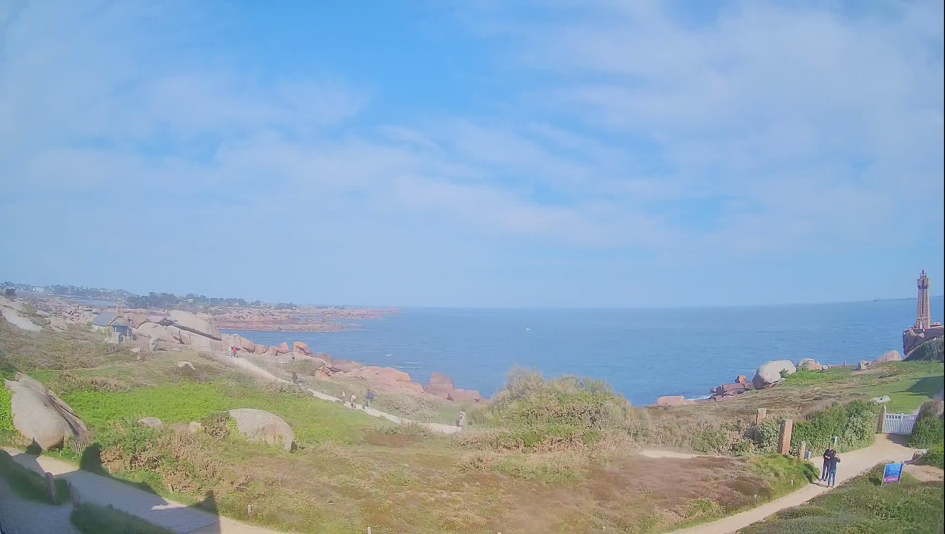 A coastal path winds through grassy terrain dotted with large pink rocks, leading to a lighthouse visible in the distance on a partly cloudy, sunny day.