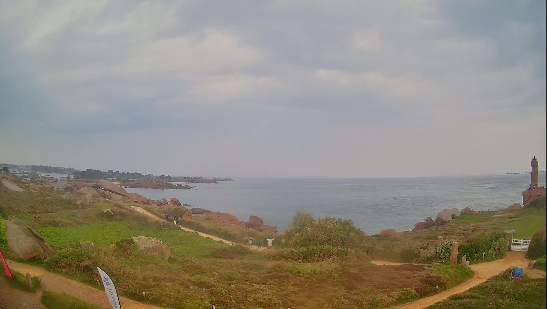 A coastal scene under an overcast sky shows a rocky shoreline with a lighthouse in the distance, and a few people walking on a path near the water.