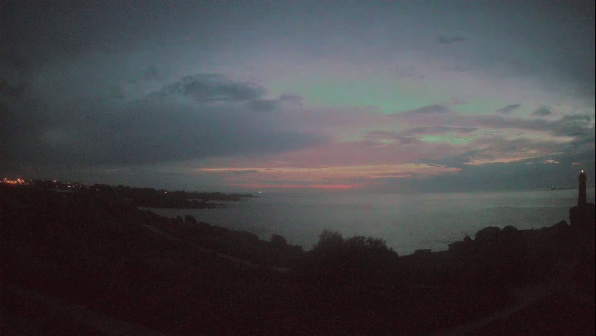 A hazy sunset over a calm ocean, seen from a rocky coastline with a distant lighthouse.