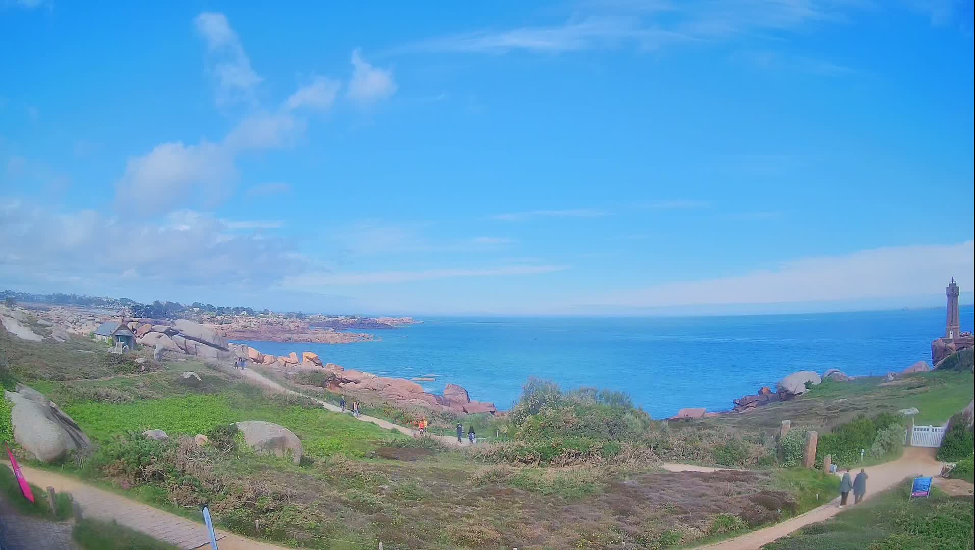 A coastal path winds through grassy terrain dotted with large rocks, leading to a blue ocean under a mostly sunny sky with a few clouds; a lighthouse stands in the distance on a rocky outcrop.