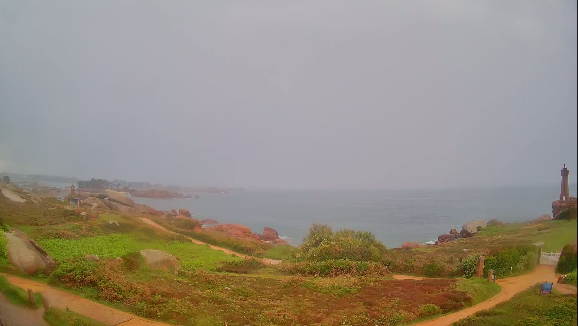 A hazy coastal scene shows a lighthouse in the distance, with a rocky shoreline and grassy paths leading to it under an overcast sky.