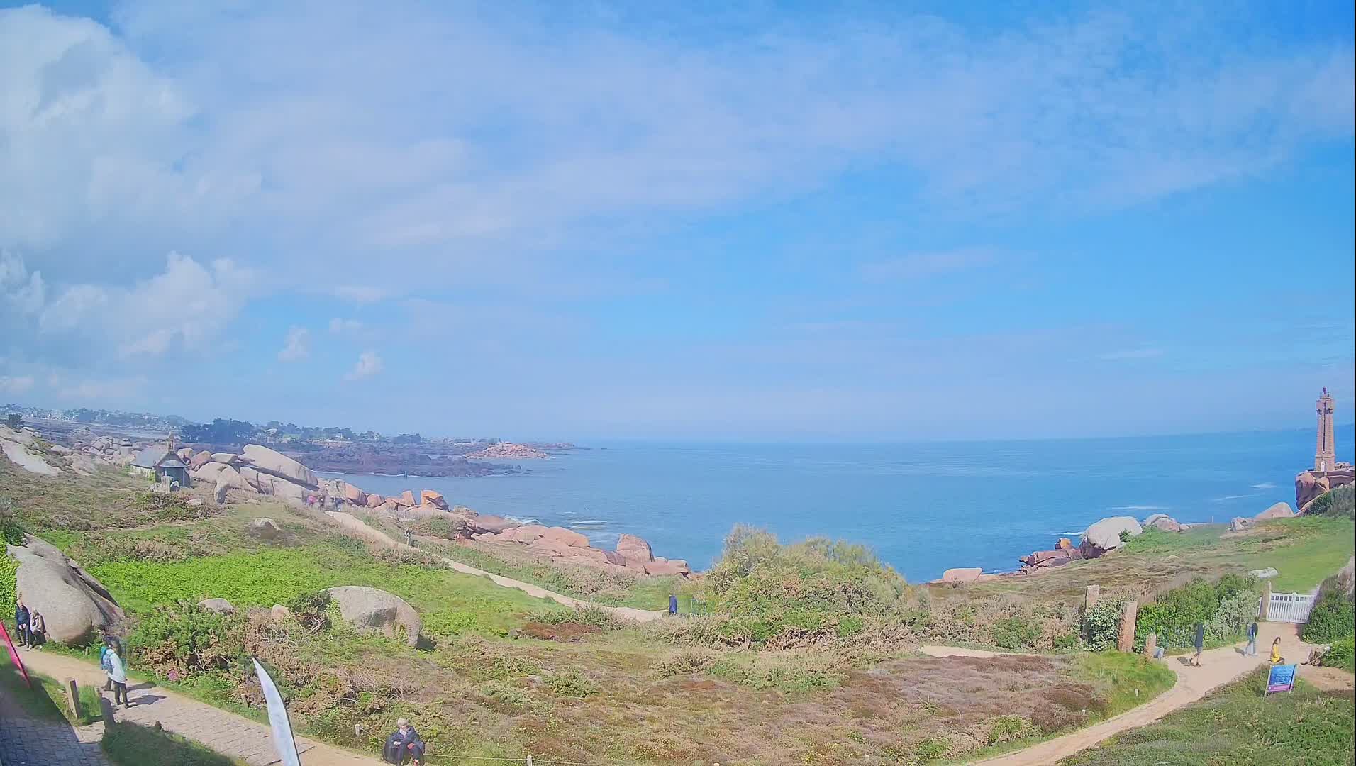 A partly cloudy sky overlooks a coastal path winding through grassy terrain dotted with large rocks, leading toward a calm ocean and a distant lighthouse.