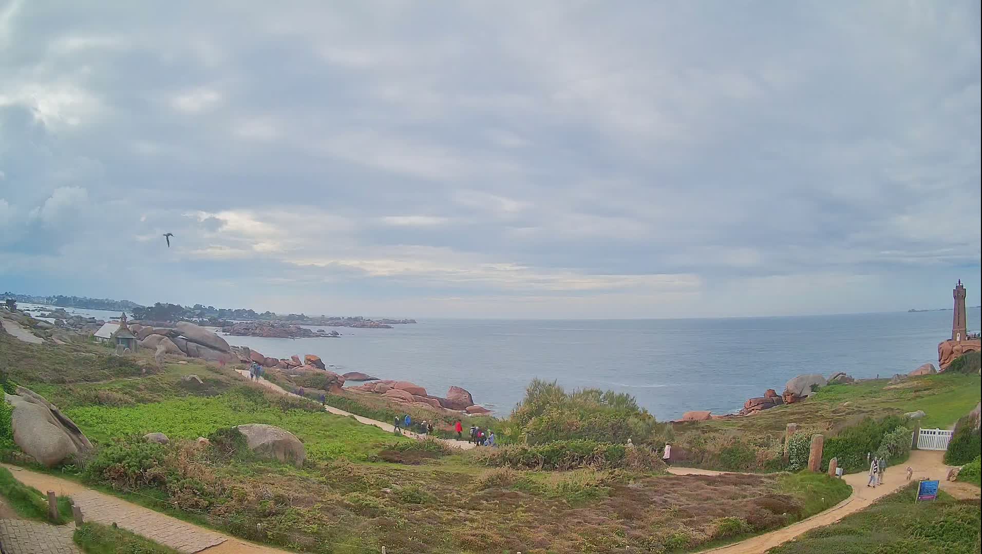 A coastal scene under a cloudy sky shows a group of people walking along a path near pink granite rocks and a lighthouse in the distance, with a calm ocean stretching to the horizon.