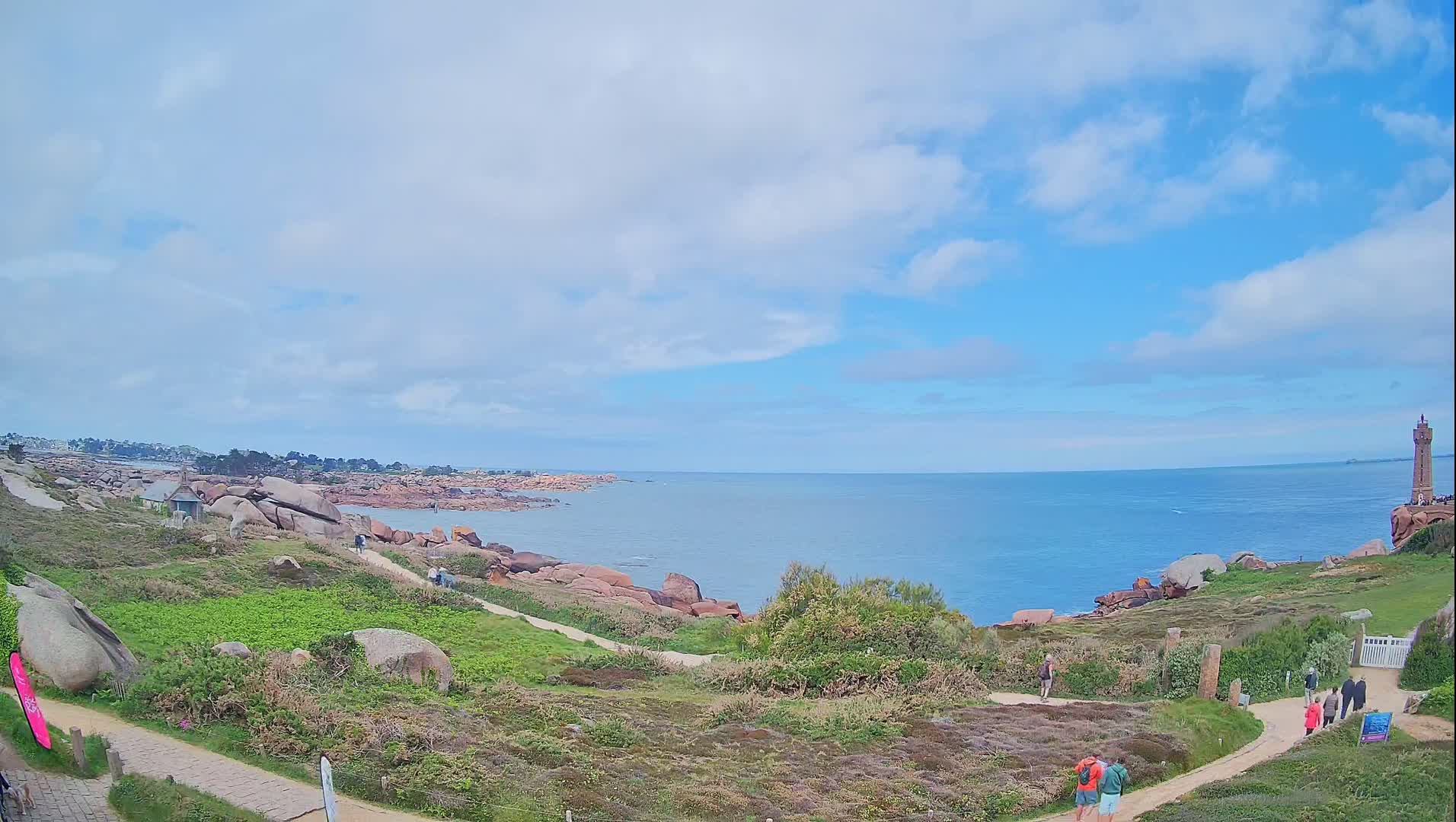 A partly cloudy day reveals a coastal scene with pink rocks, green vegetation, a lighthouse in the distance, and a few people walking along a path overlooking a calm ocean.