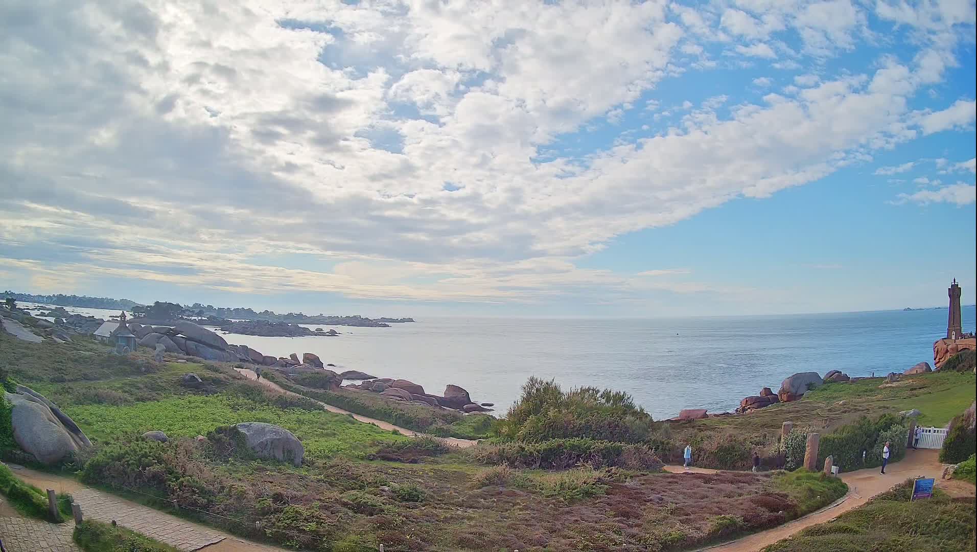 A coastal scene under a partly cloudy sky shows a rocky shoreline with walking paths, green vegetation, and a lighthouse in the distance.
