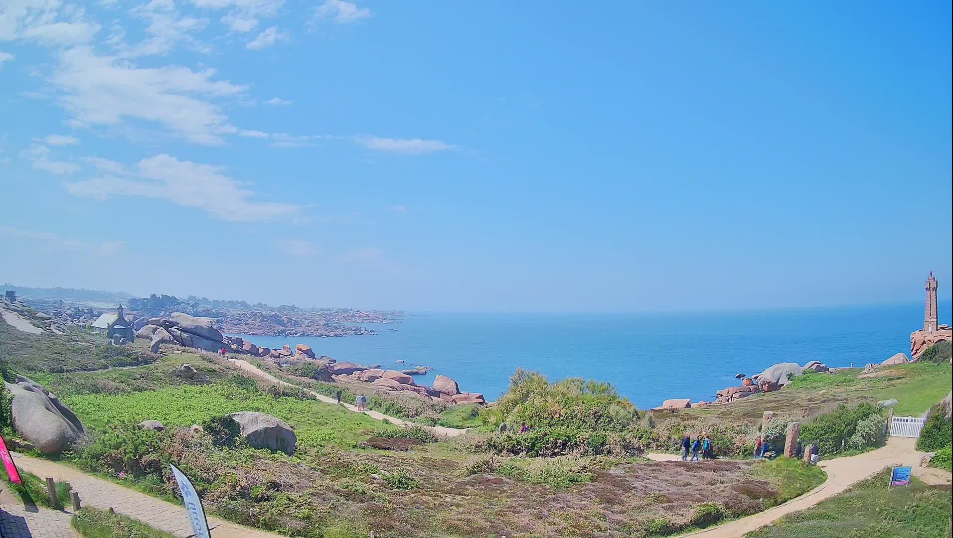 A coastal path winds through a rocky, grassy landscape overlooking a calm blue ocean under a mostly sunny sky.