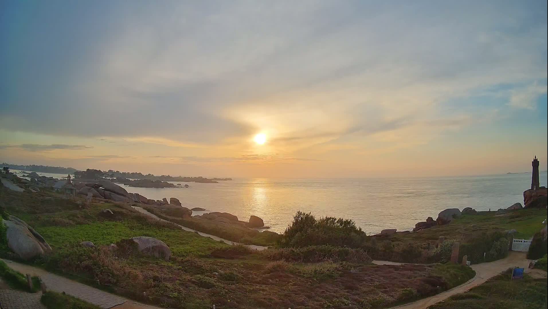 A coastal sunset scene shows a calm ocean reflecting the golden light, with rocky shores, low-lying vegetation, and a distant lighthouse.
