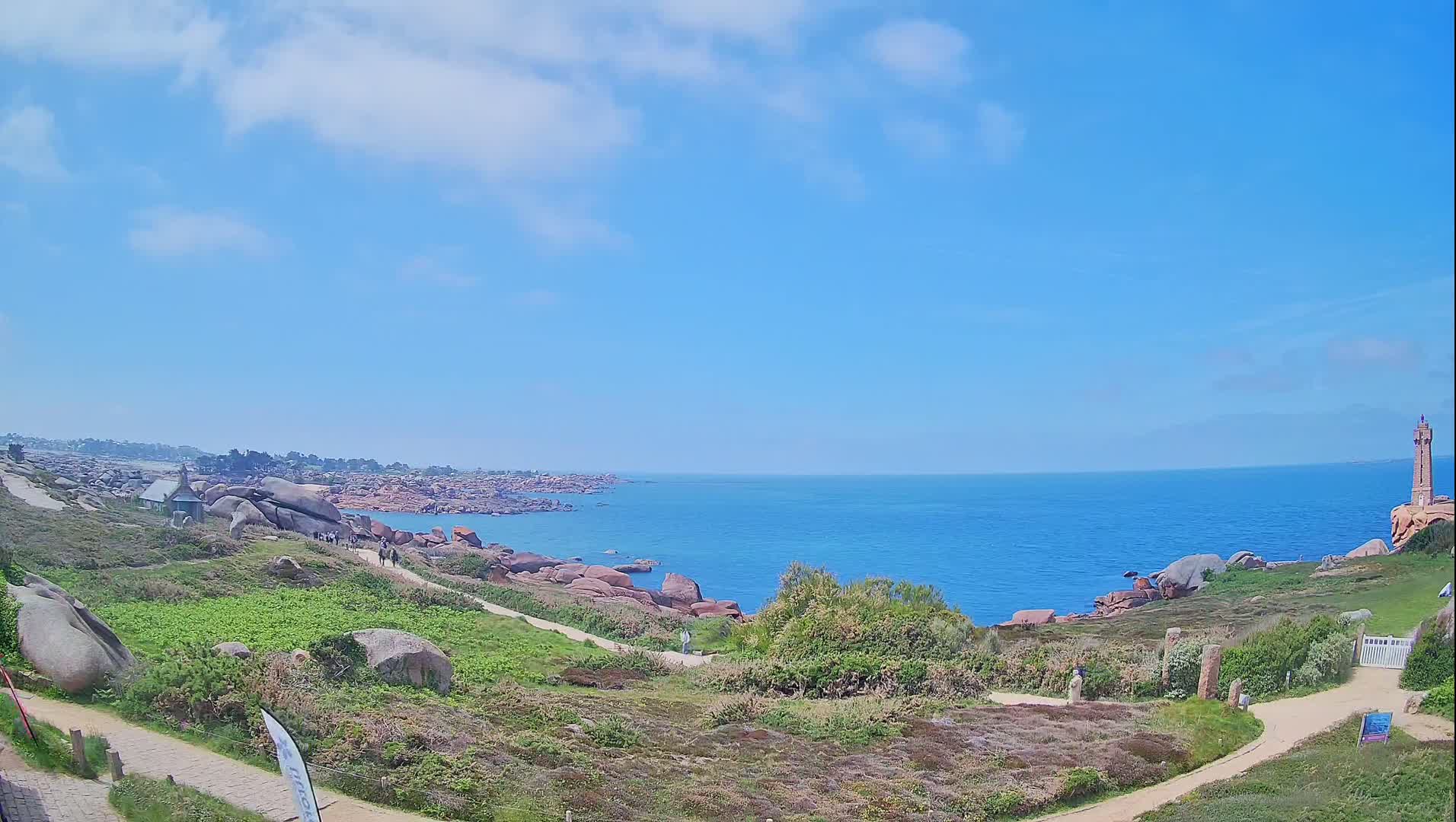 A coastal path winds through grassy terrain dotted with large rocks, overlooking a calm blue ocean under a mostly sunny sky, with a lighthouse visible in the distance.