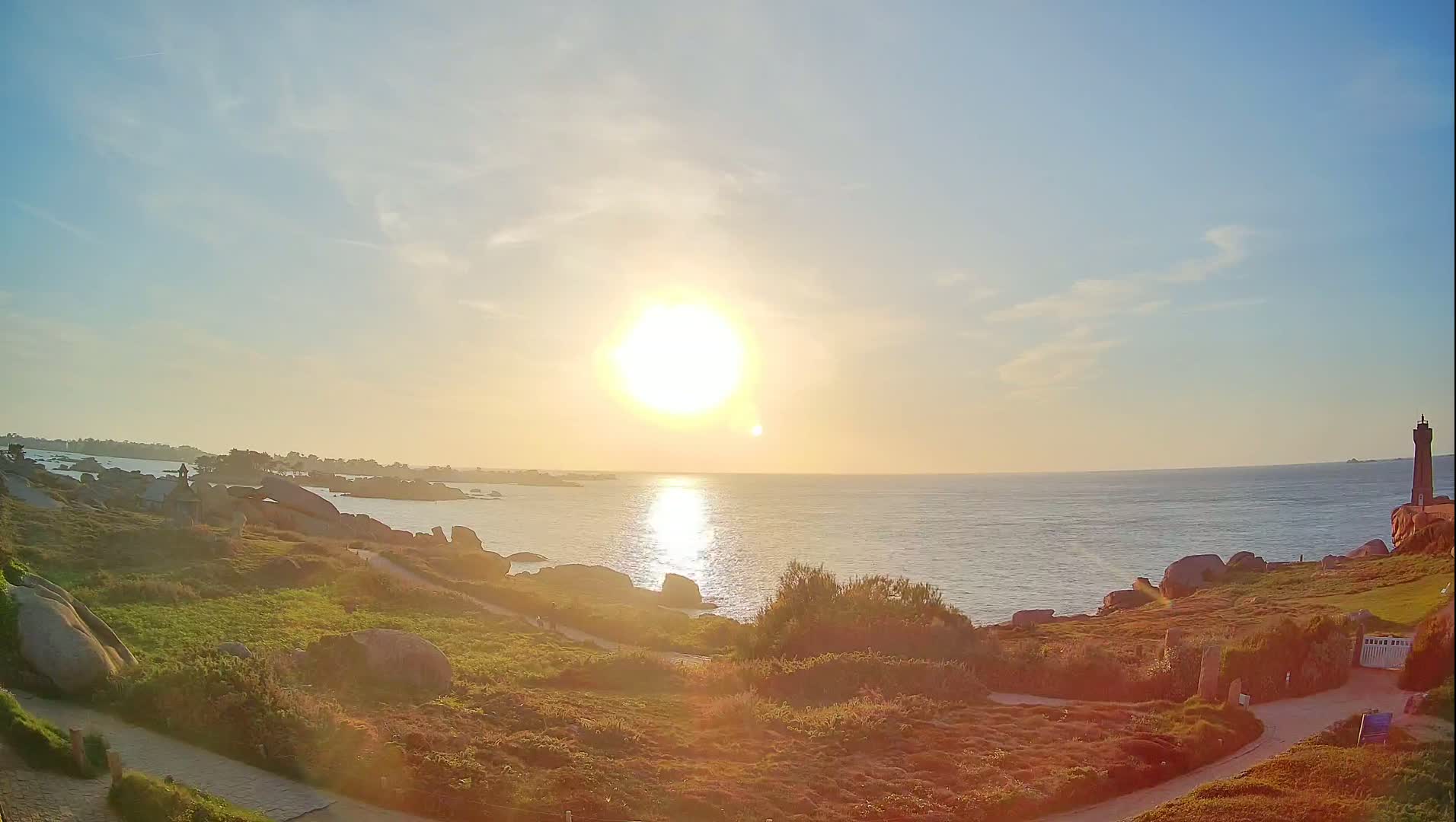 A coastal scene at sunset shows a lighthouse in the distance, rocky terrain, and a calm ocean reflecting the sun's light.