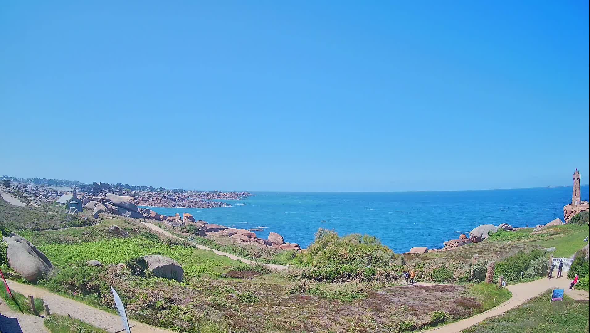 A coastal scene on a sunny day shows a lighthouse in the distance, a rocky shoreline with walking paths, and sparse vegetation.