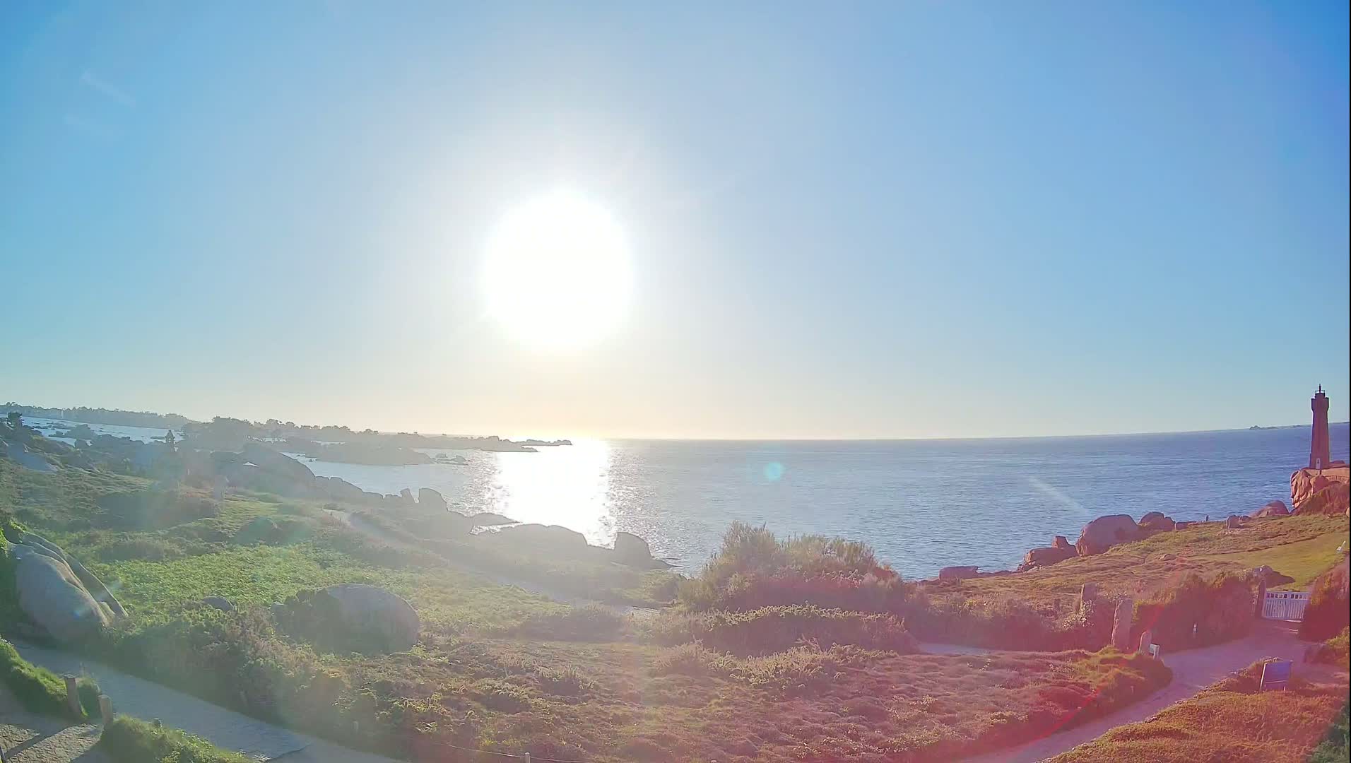 A bright, sunny day at a coastal area with a lighthouse visible in the distance, featuring rocky terrain and low-lying vegetation.