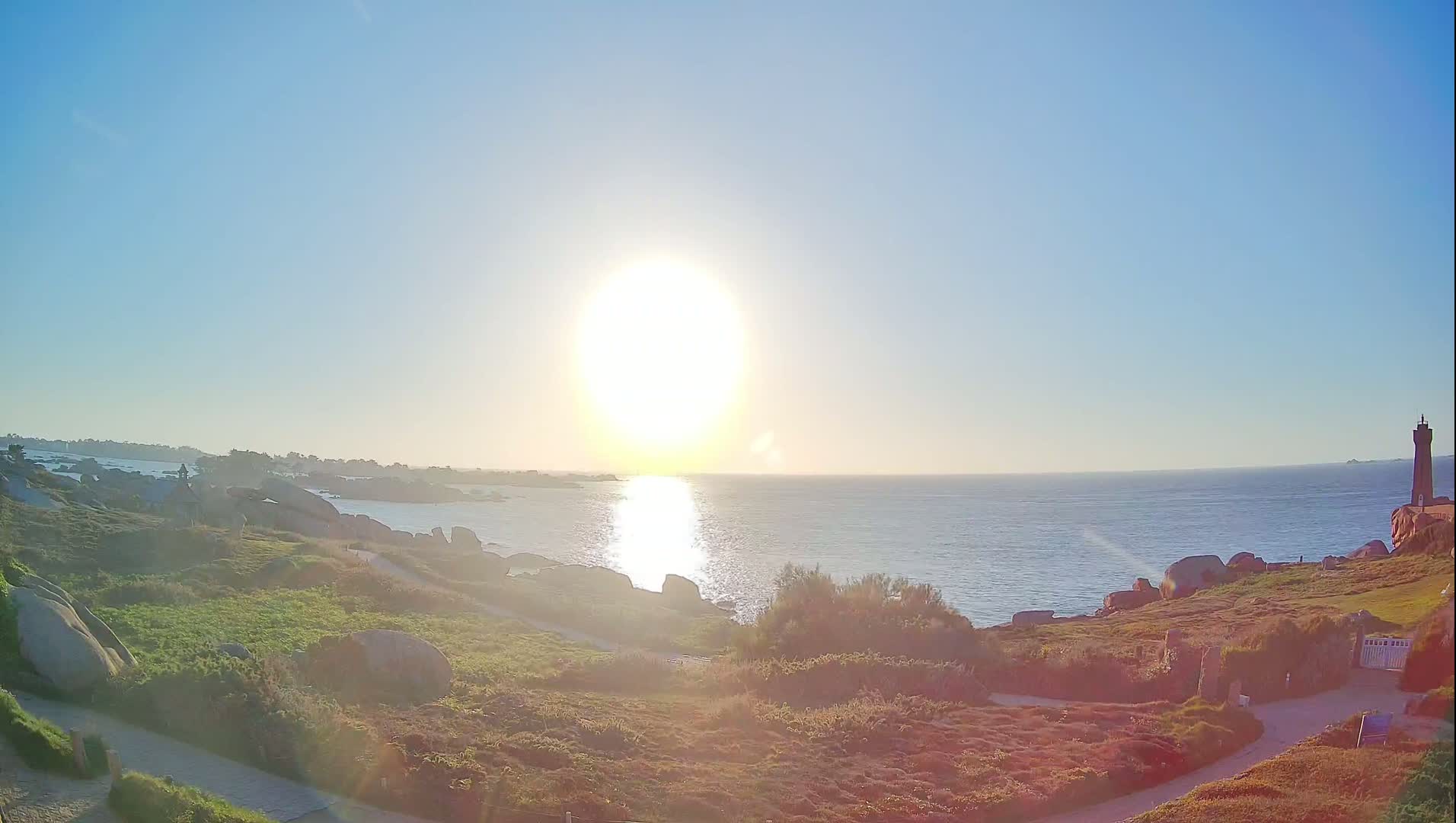 A bright sun shines over a rocky coastline and calm ocean, with a lighthouse visible in the distance.