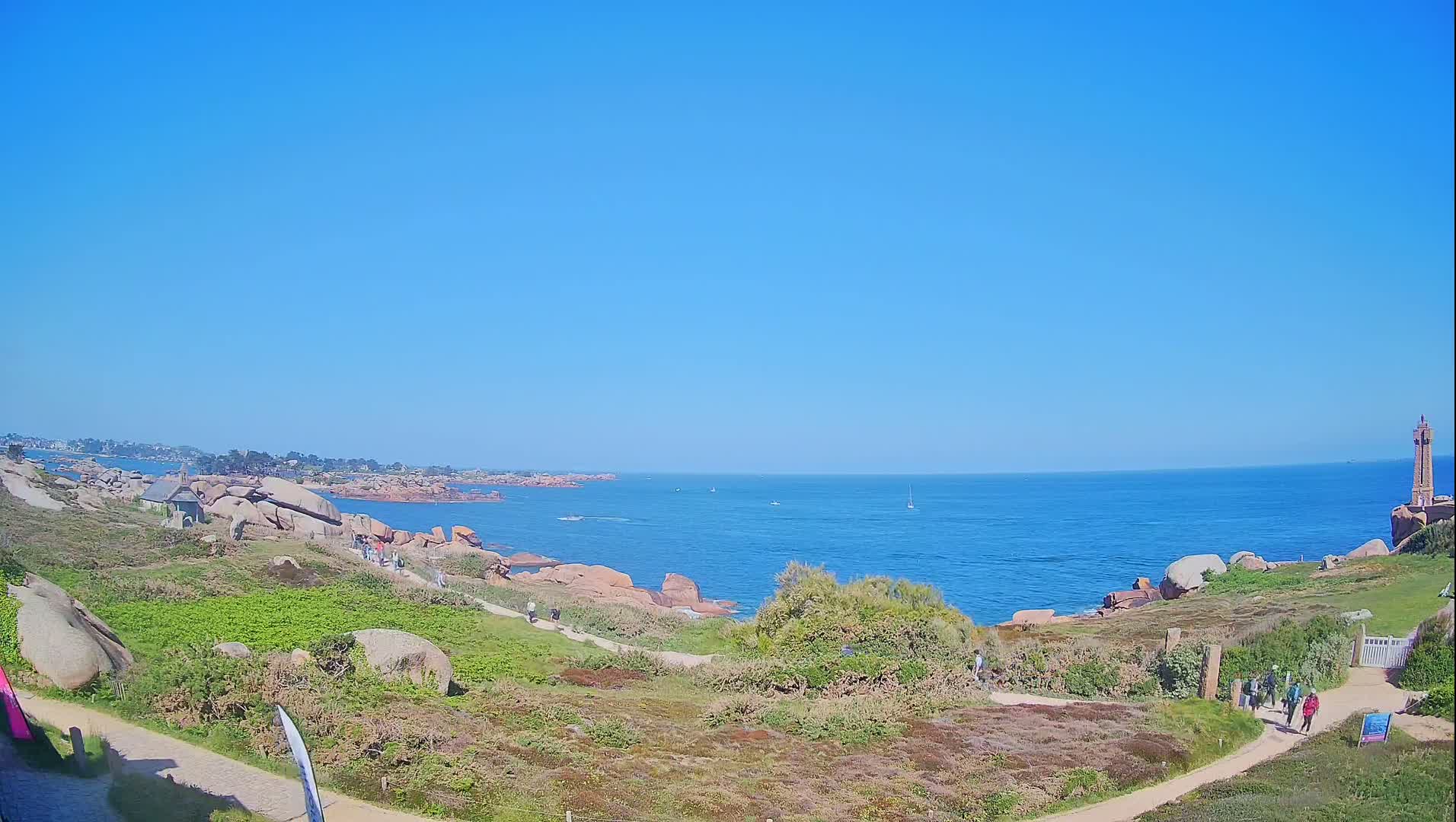 A coastal path winds through grassy hills dotted with pink rocks toward a lighthouse on a clear, sunny day, overlooking a calm blue ocean.