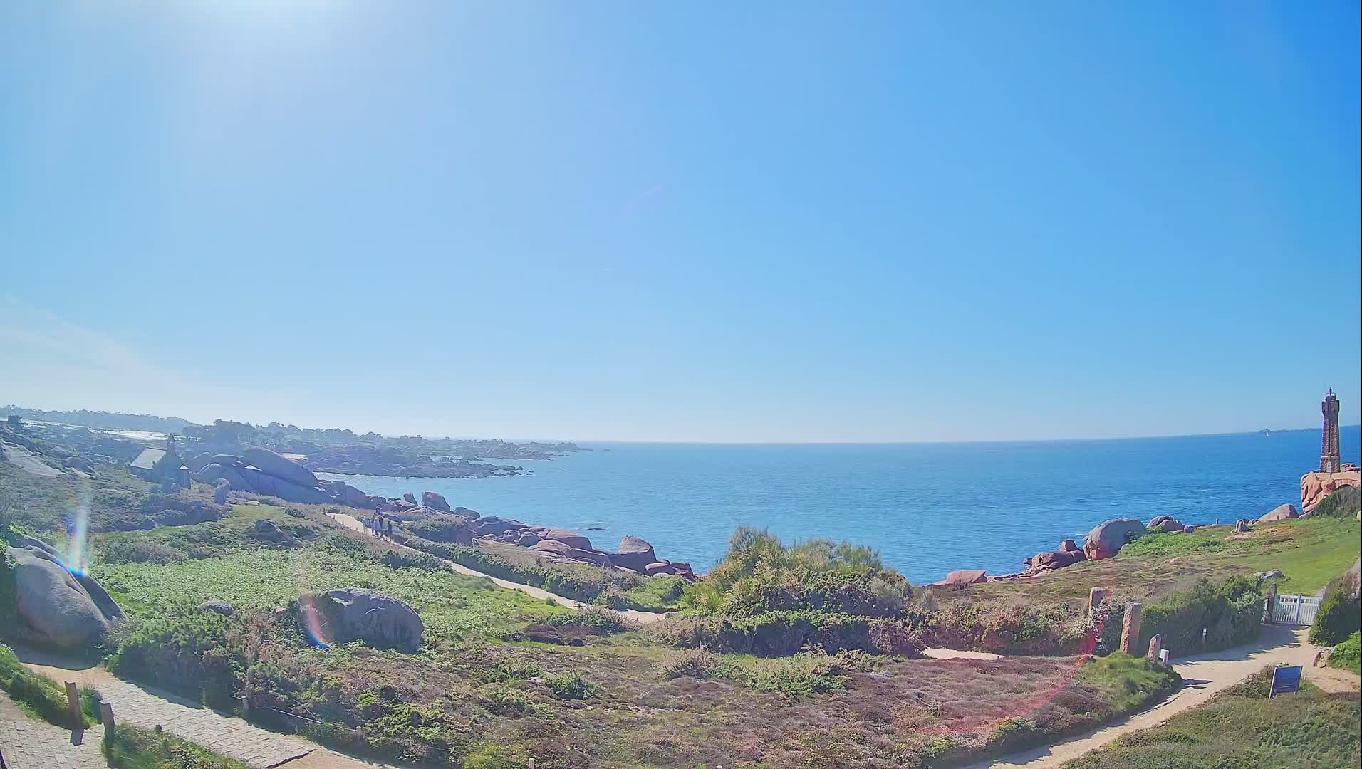 A coastal path winds through a rocky, grassy landscape overlooking a calm, blue ocean under a bright, sunny sky, with a lighthouse visible in the distance.