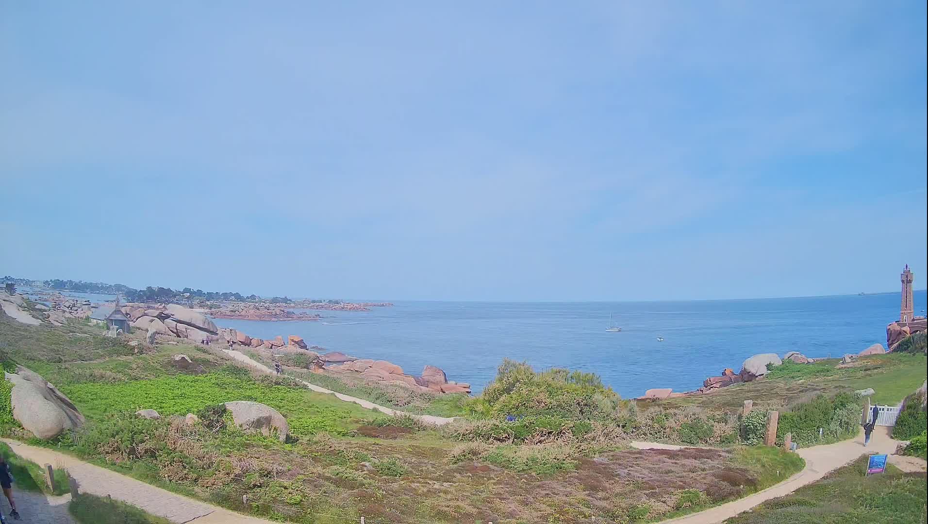A coastal scene under a clear, light blue sky shows a rocky shoreline, a calm ocean with a few boats, and a lighthouse in the distance.