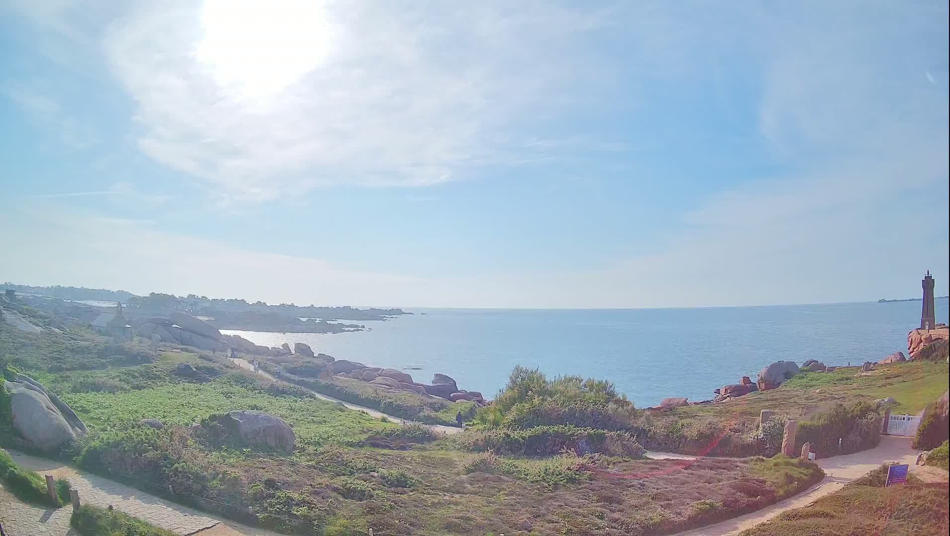 A sunny day reveals a coastal scene with a lighthouse in the distance, a rocky shoreline, and a path winding through low-lying vegetation.