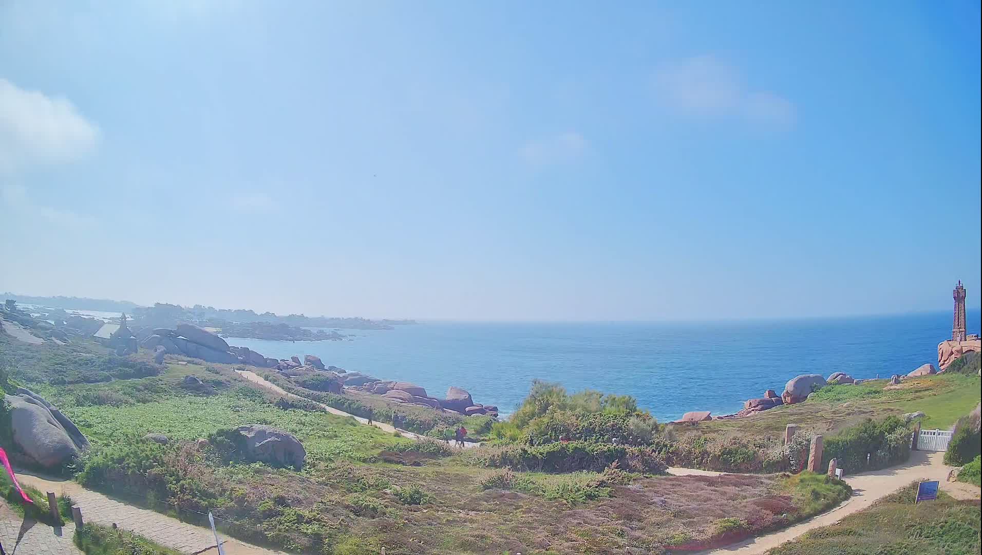 A coastal path winds through a rocky, grassy landscape overlooking a calm, blue ocean under a clear, sunny sky, with a lighthouse visible in the distance.