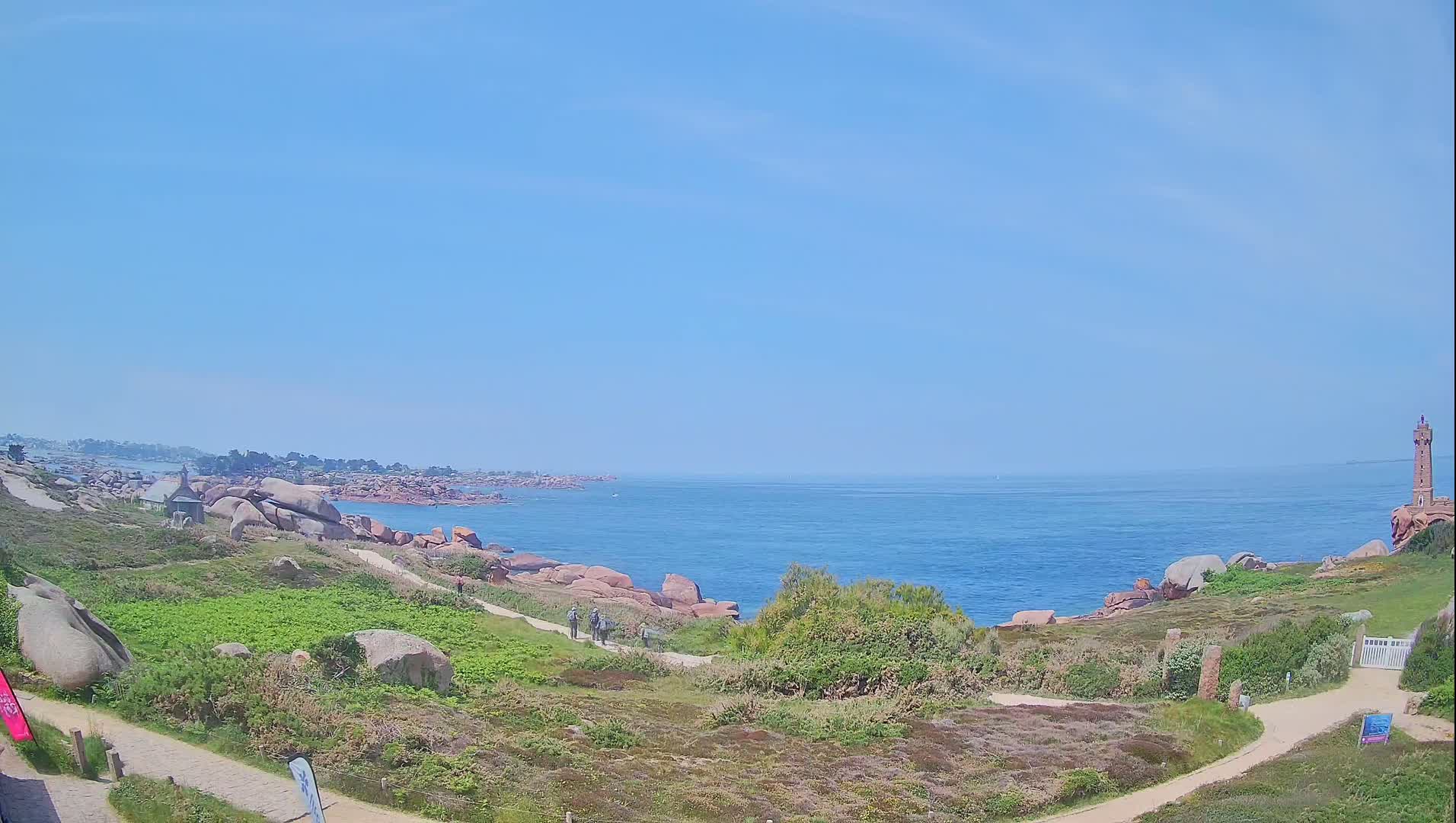 A coastal path winds through grassy terrain dotted with pink granite boulders, leading to a calm ocean under a clear blue sky, with a lighthouse visible in the distance.