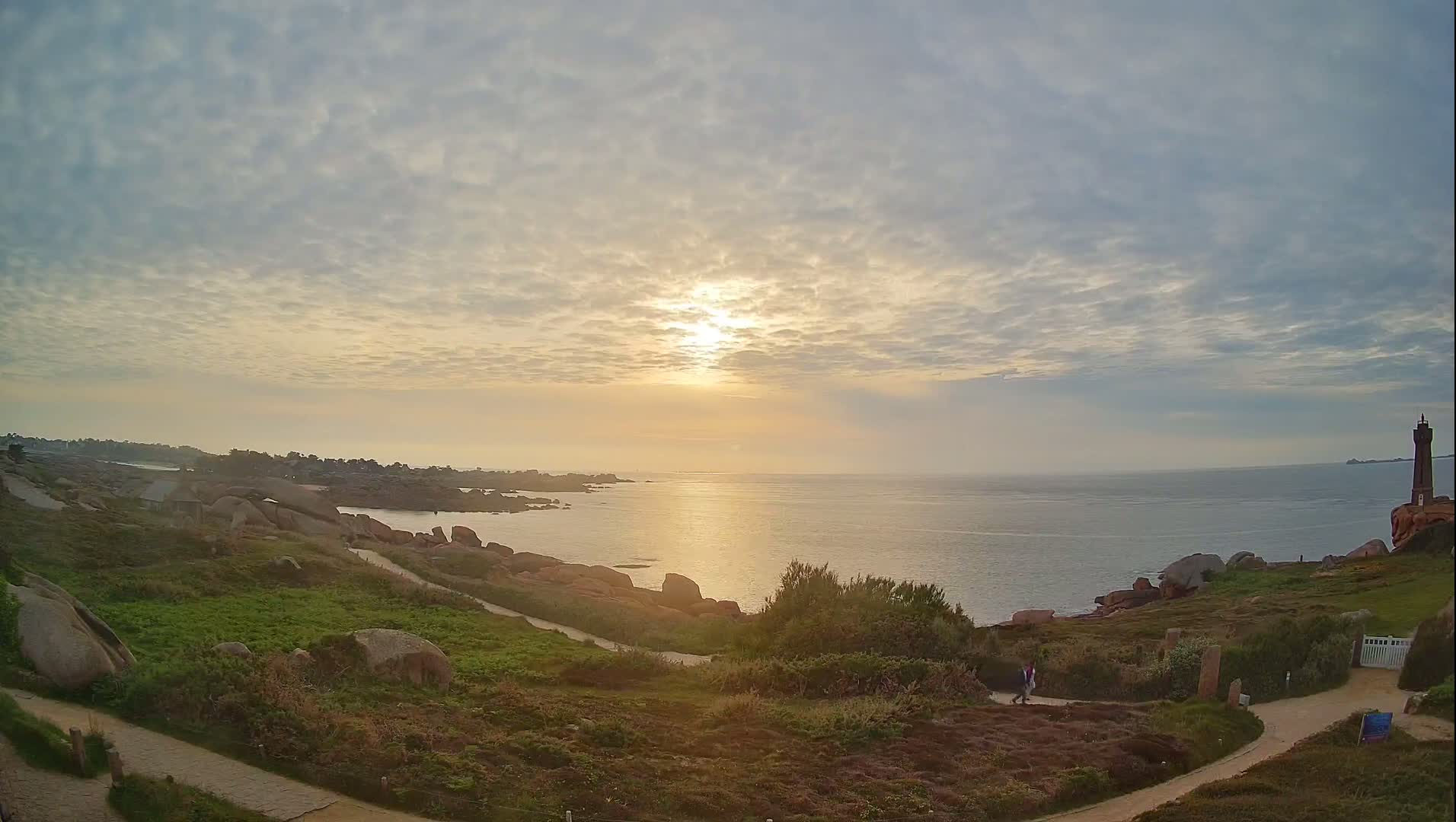 A coastal scene at sunset under a partly cloudy sky shows a calm sea, rocky shoreline with grassy areas, and a lighthouse in the distance.