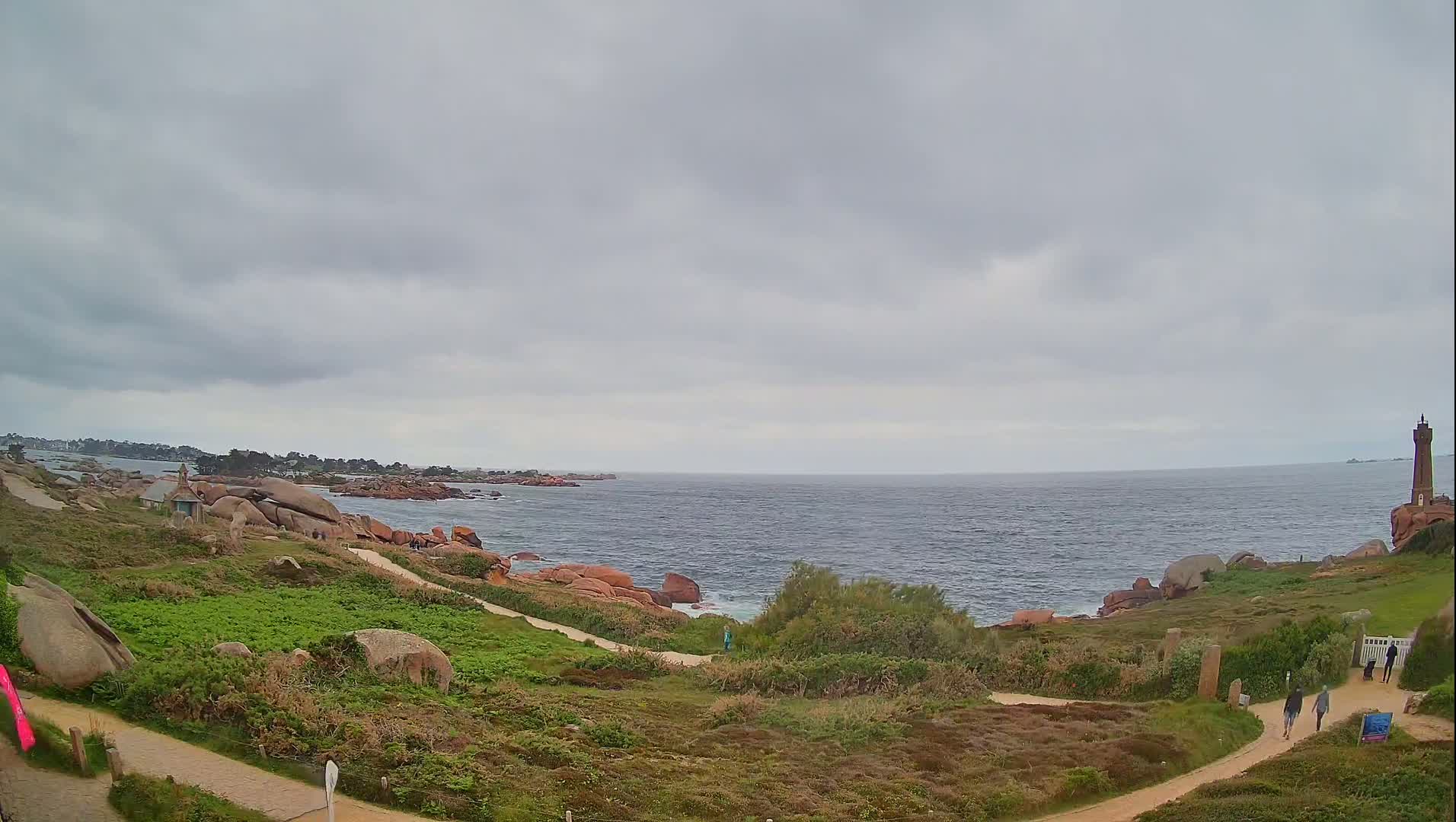 A coastal path winds through low-lying vegetation towards a lighthouse on a rocky outcrop overlooking a calm sea under an overcast sky.