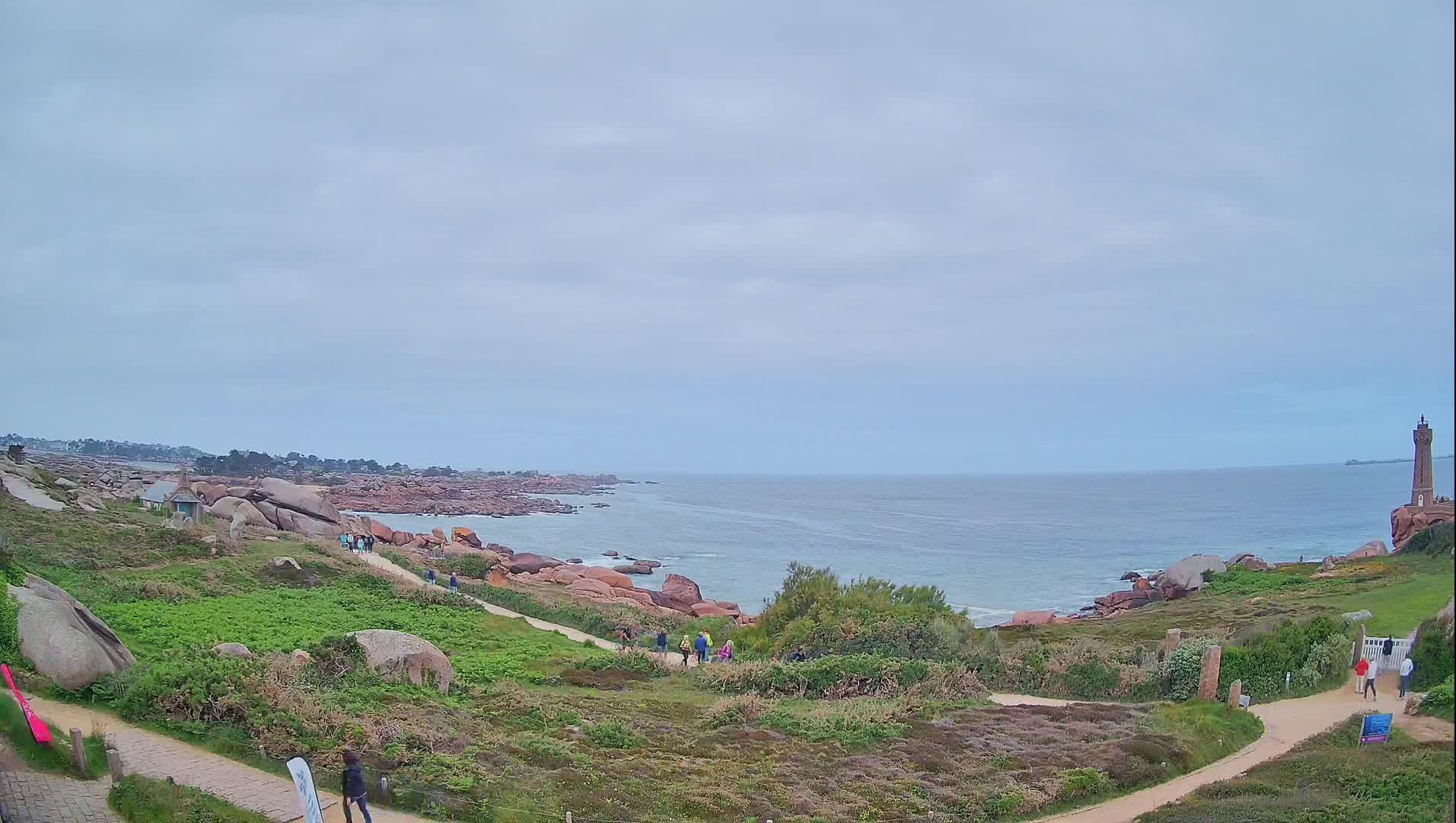 A coastal path winds through green vegetation alongside a rocky shoreline, with a lighthouse visible in the distance under a cloudy sky.