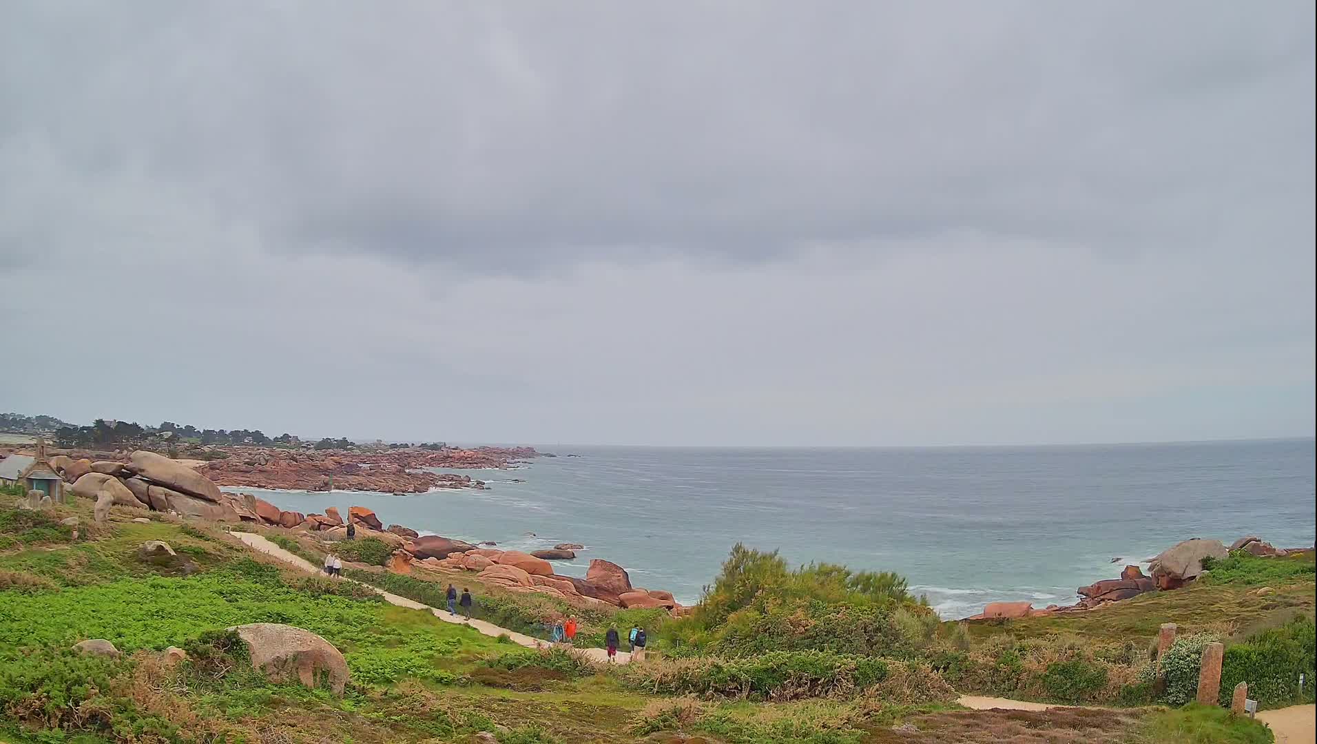 A coastal path winds through green vegetation towards a calm sea under a cloudy sky, with several people walking along the path.