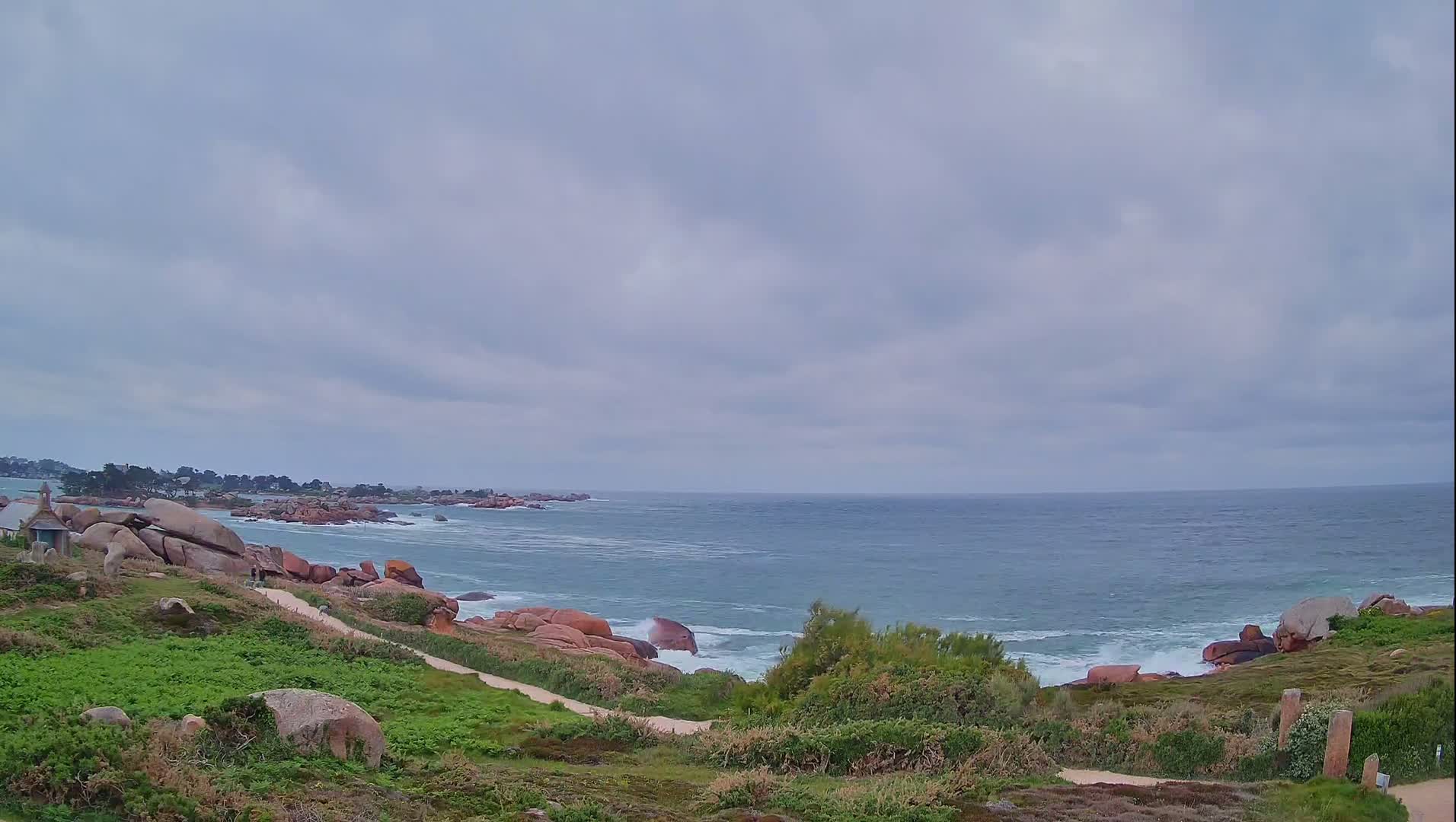 A coastal path winds through green vegetation alongside pink rocks overlooking a blue ocean with small waves under a cloudy sky.
