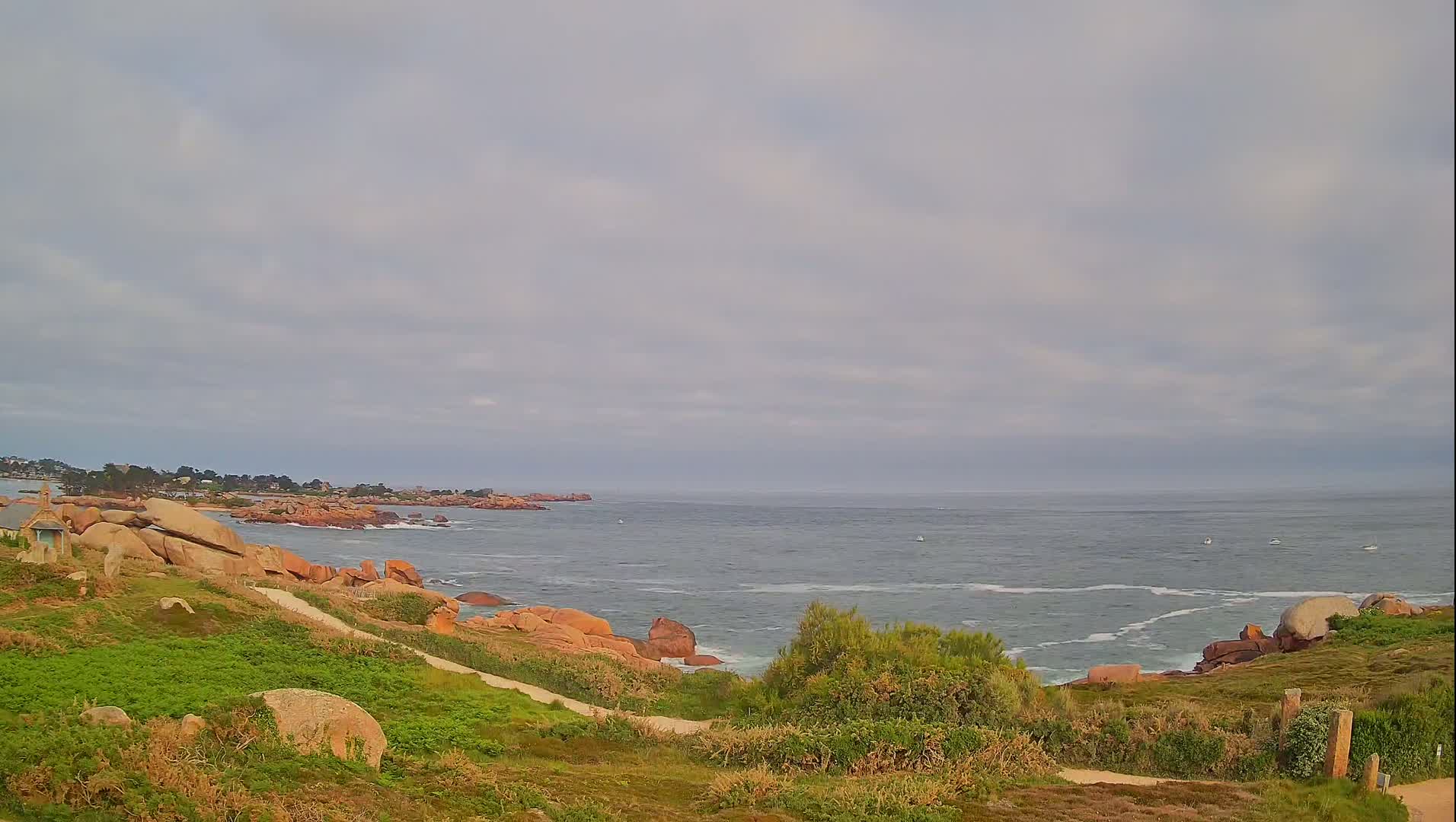A coastal scene under a cloudy sky shows a rocky shoreline with green vegetation, a calm sea with a few small boats, and a small town visible in the distance.