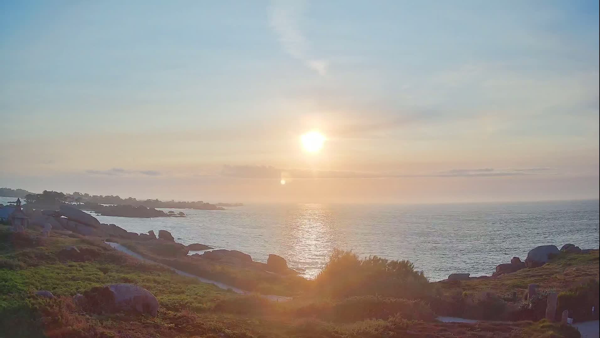 A setting sun casts a warm glow over a rocky coastline and calm ocean.