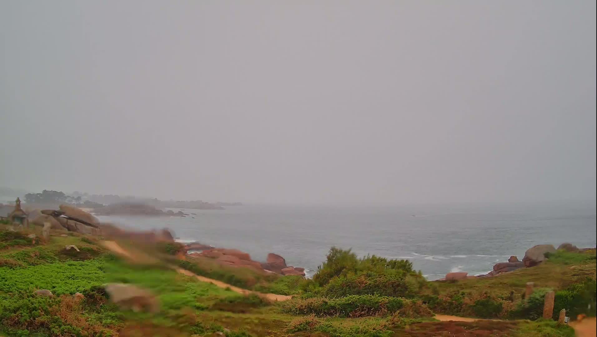 A hazy, overcast day reveals a rocky coastline with green vegetation and a calm ocean extending to the horizon.