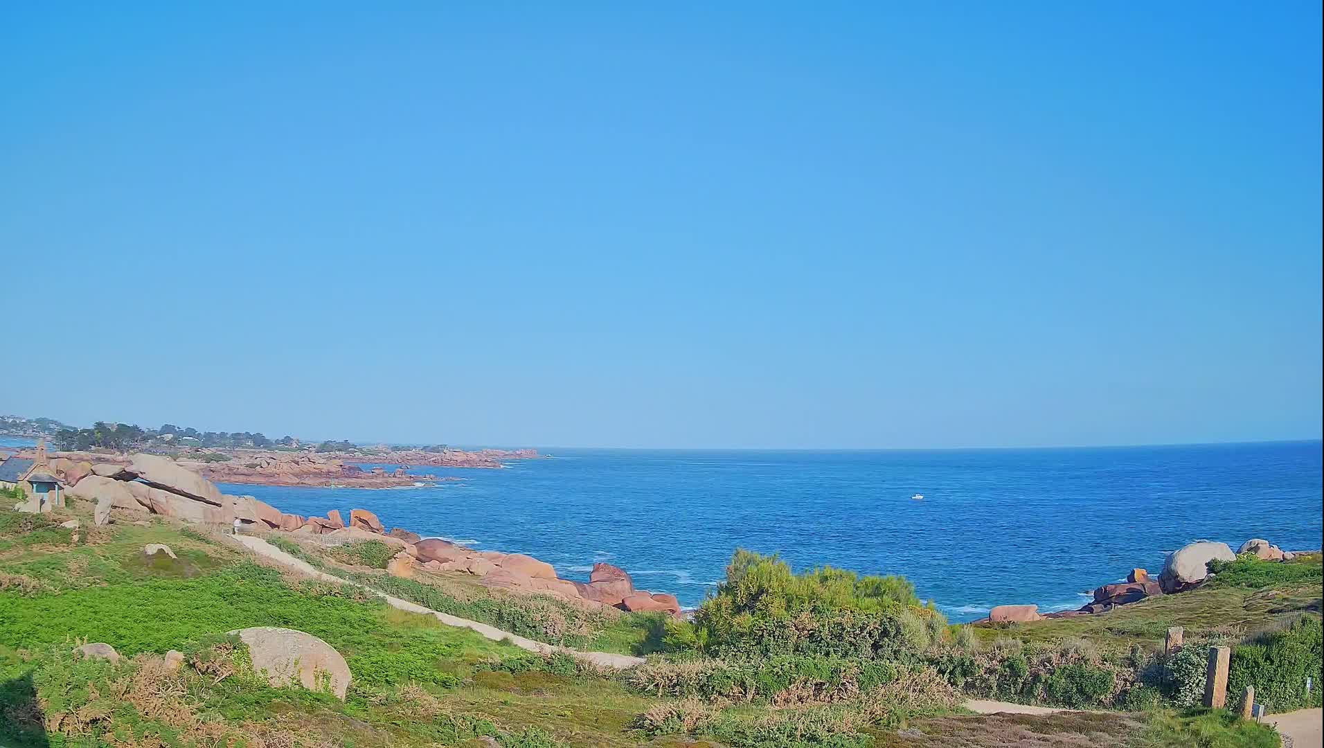 A clear blue sky overlooks a rocky coastline with green vegetation and a calm, blue ocean in the distance.