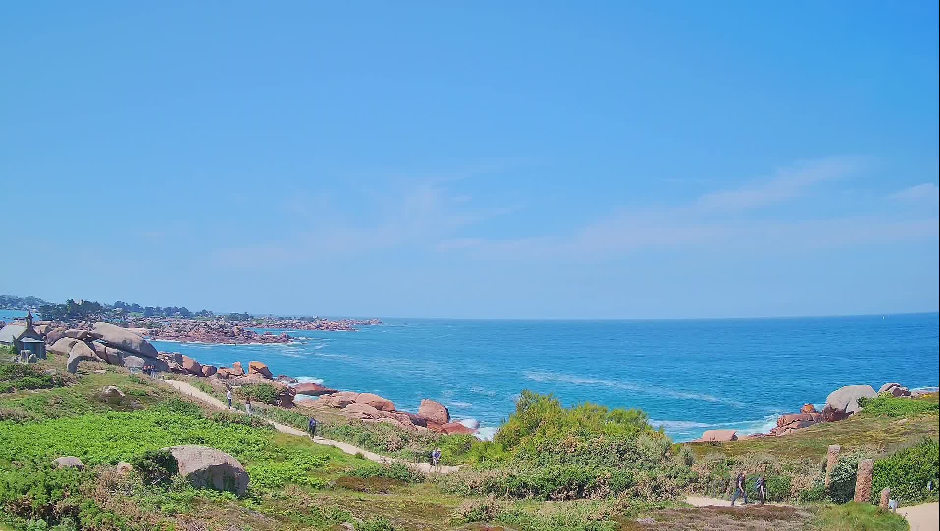 A coastal path winds through green vegetation alongside a rocky shoreline, overlooking a calm, blue ocean under a clear, sunny sky.