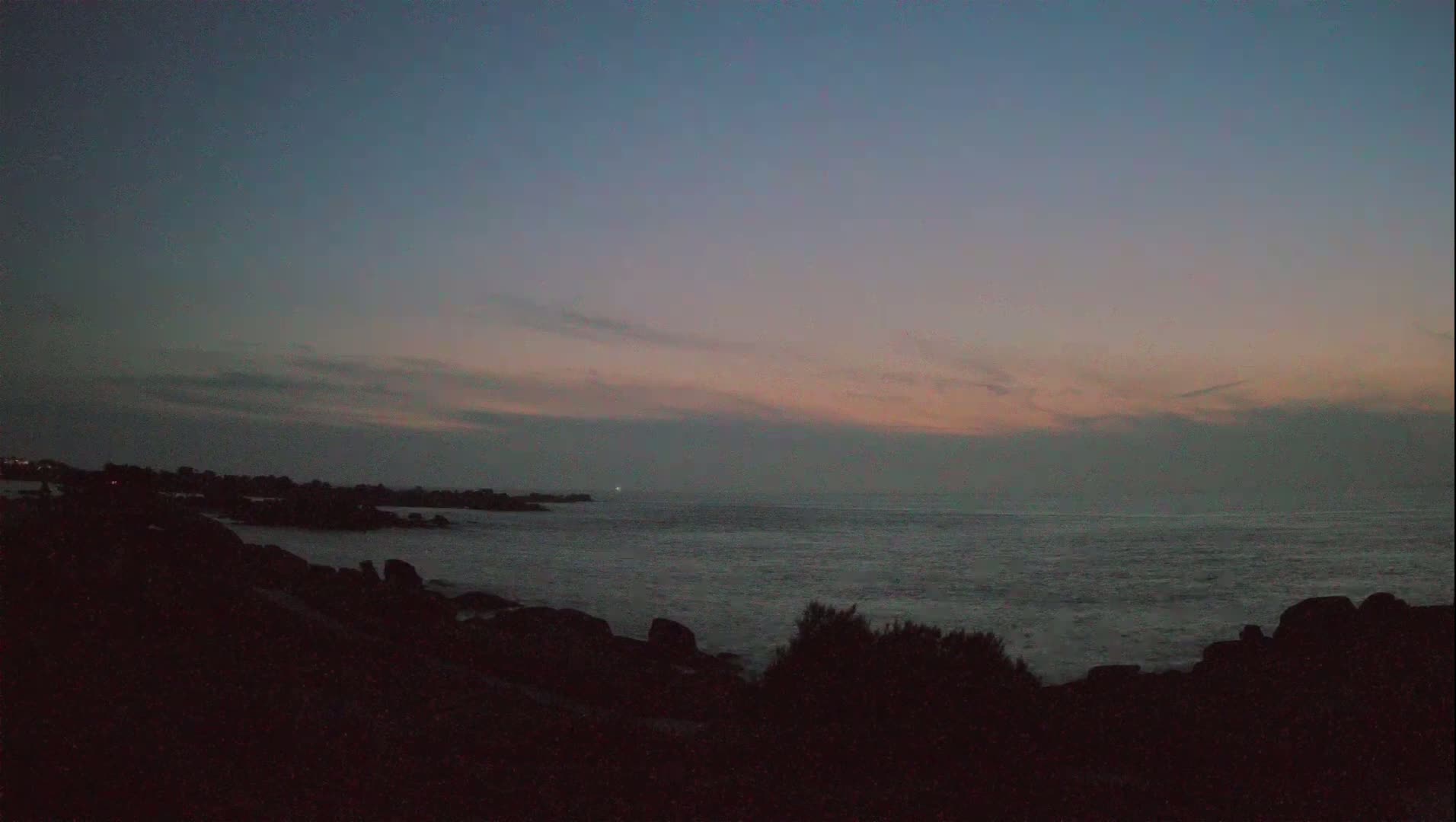 A dark, rocky shoreline meets a calm sea under a dusky, pink-tinged twilight sky.