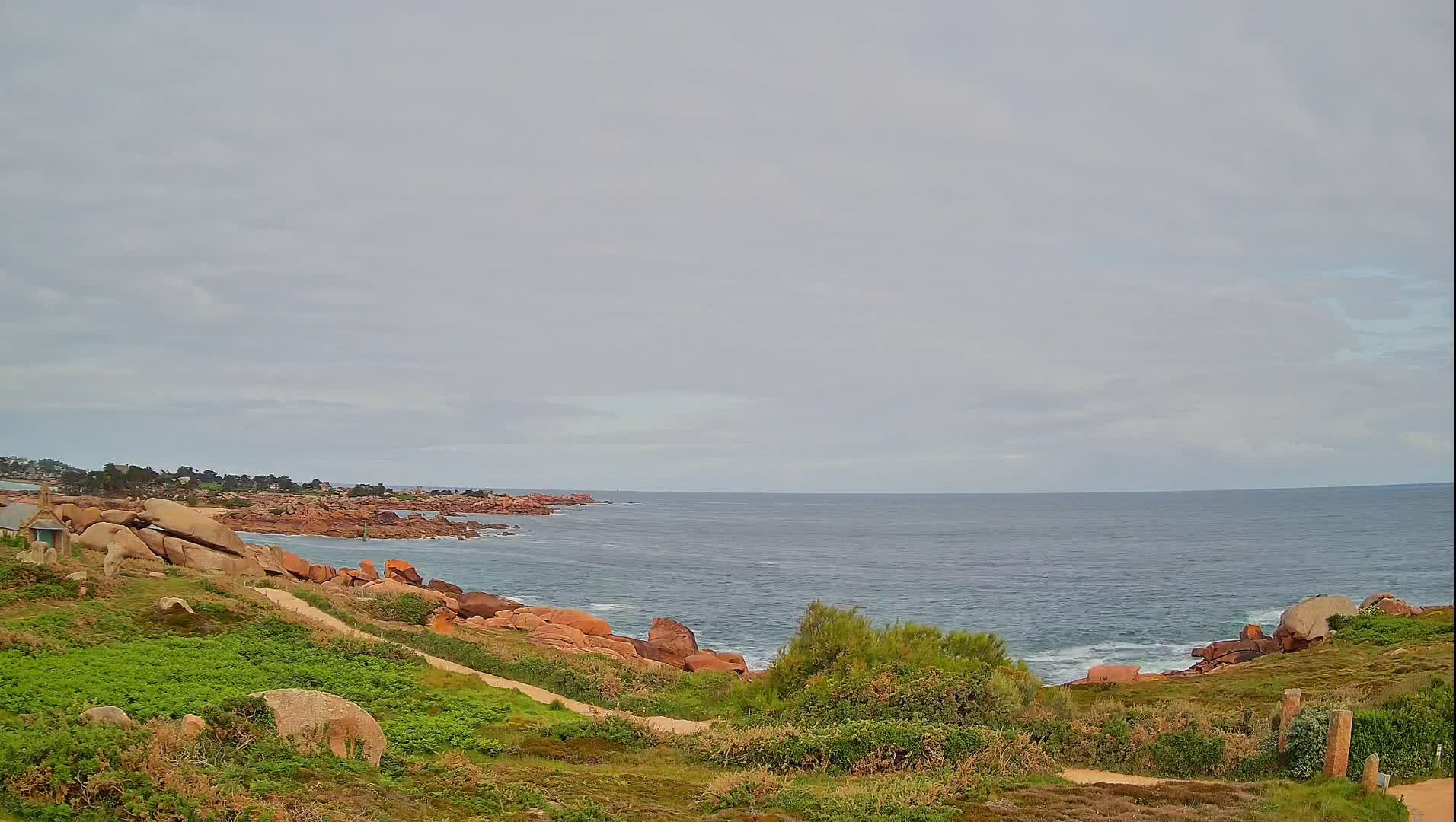 A rocky coastline with green vegetation overlooks a calm, gray ocean under a cloudy sky.