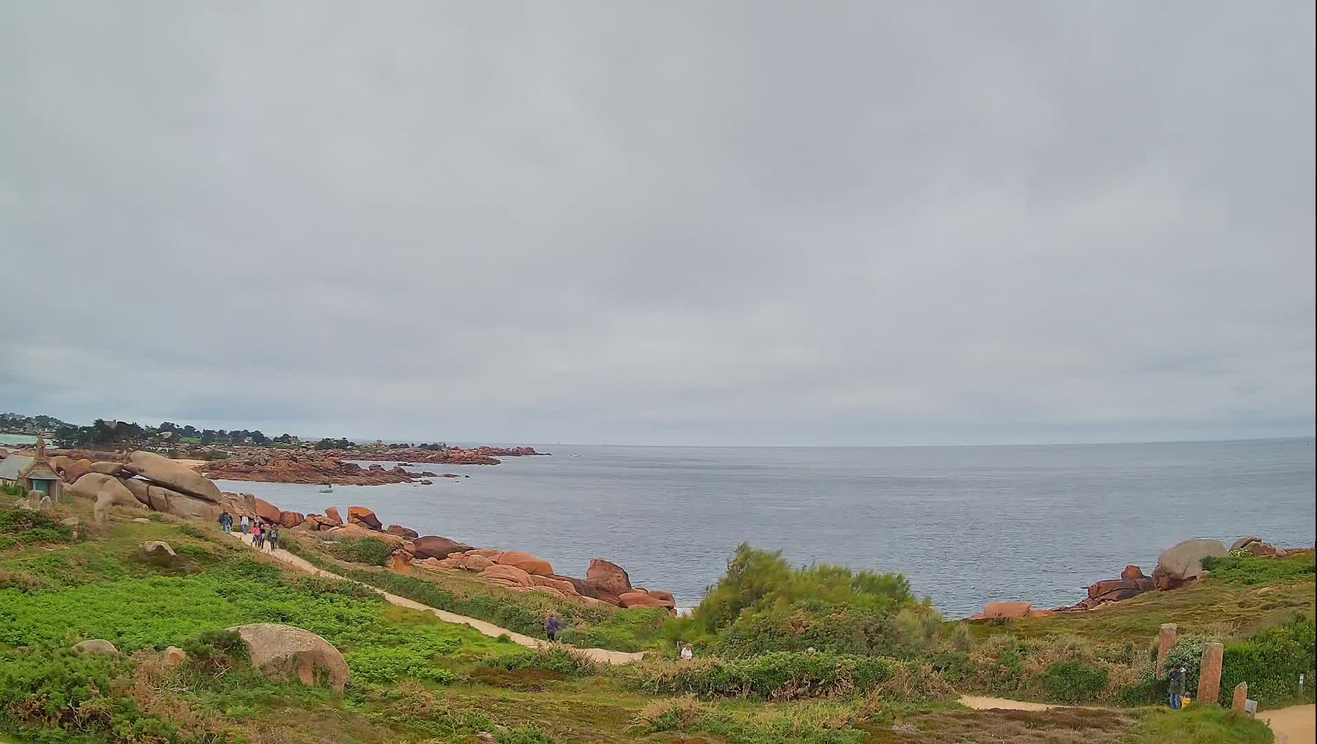 A coastal path winds through green vegetation past pink granite boulders toward a calm, gray-skied ocean.