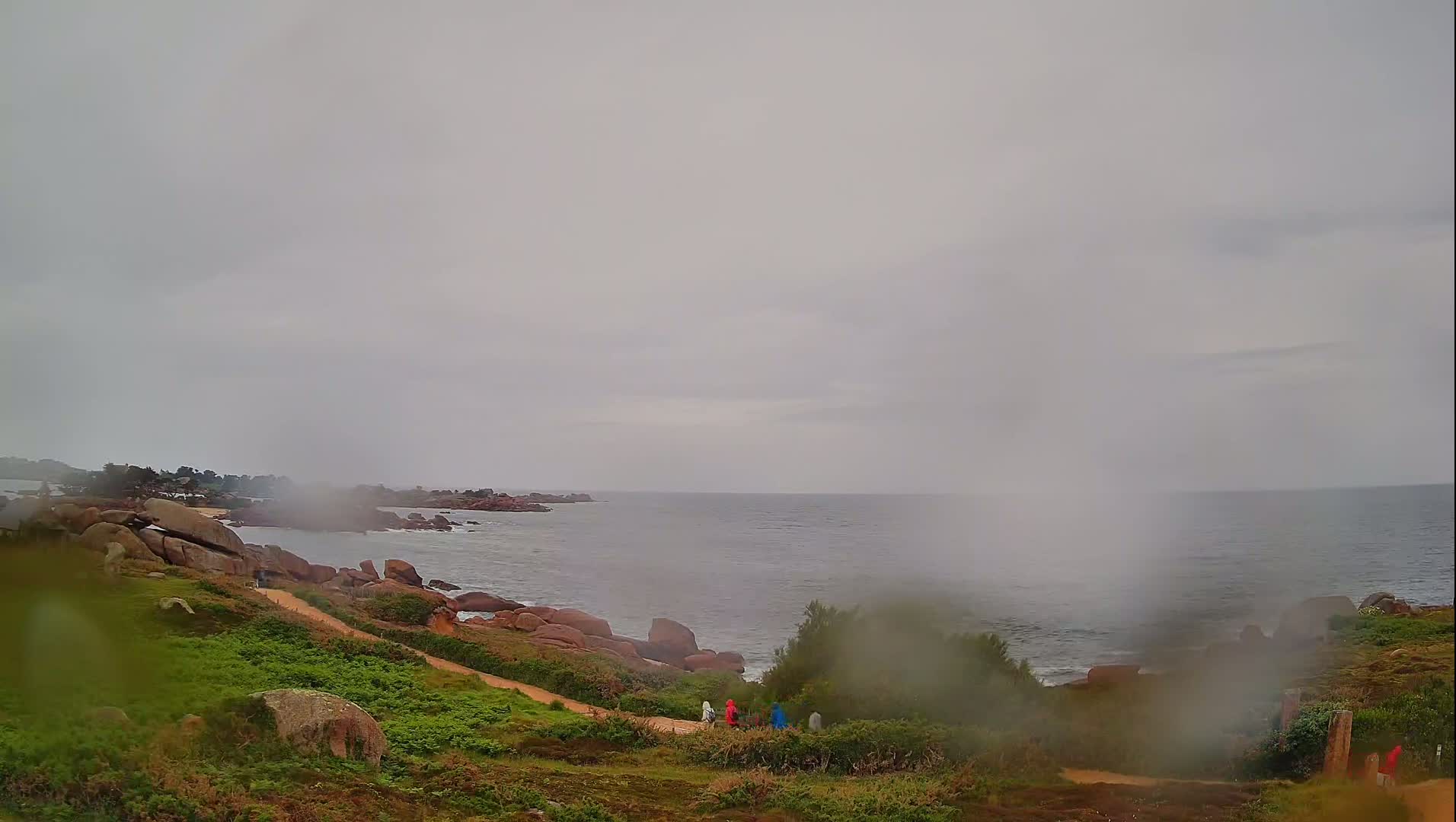 A coastal path winds through grassy terrain toward a rocky shoreline and a gray, overcast sea, with several people walking along the path in the rain.