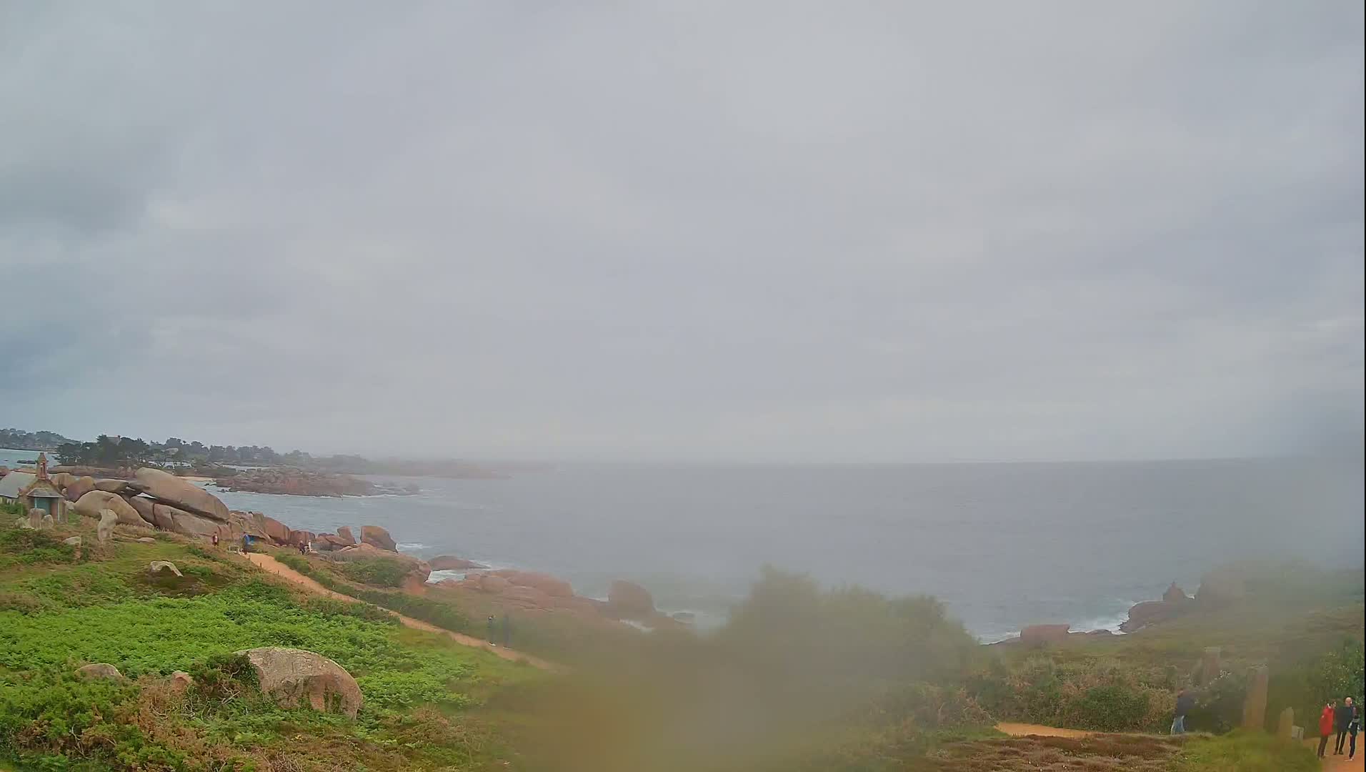 A foggy coastal scene shows a path winding down a grassy hillside toward a rocky shoreline and a calm sea under a gray sky.