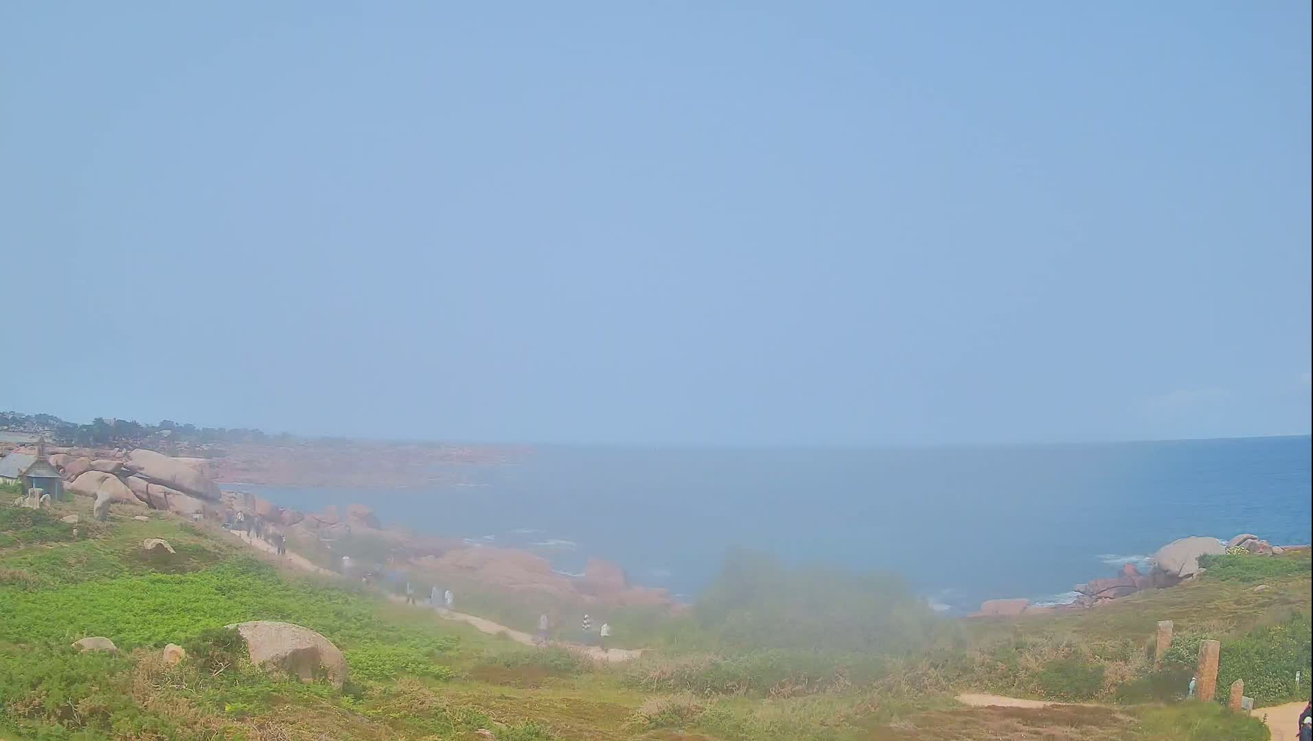 A hazy coastal scene shows a path winding down a grassy hillside dotted with large rocks toward a calm sea.