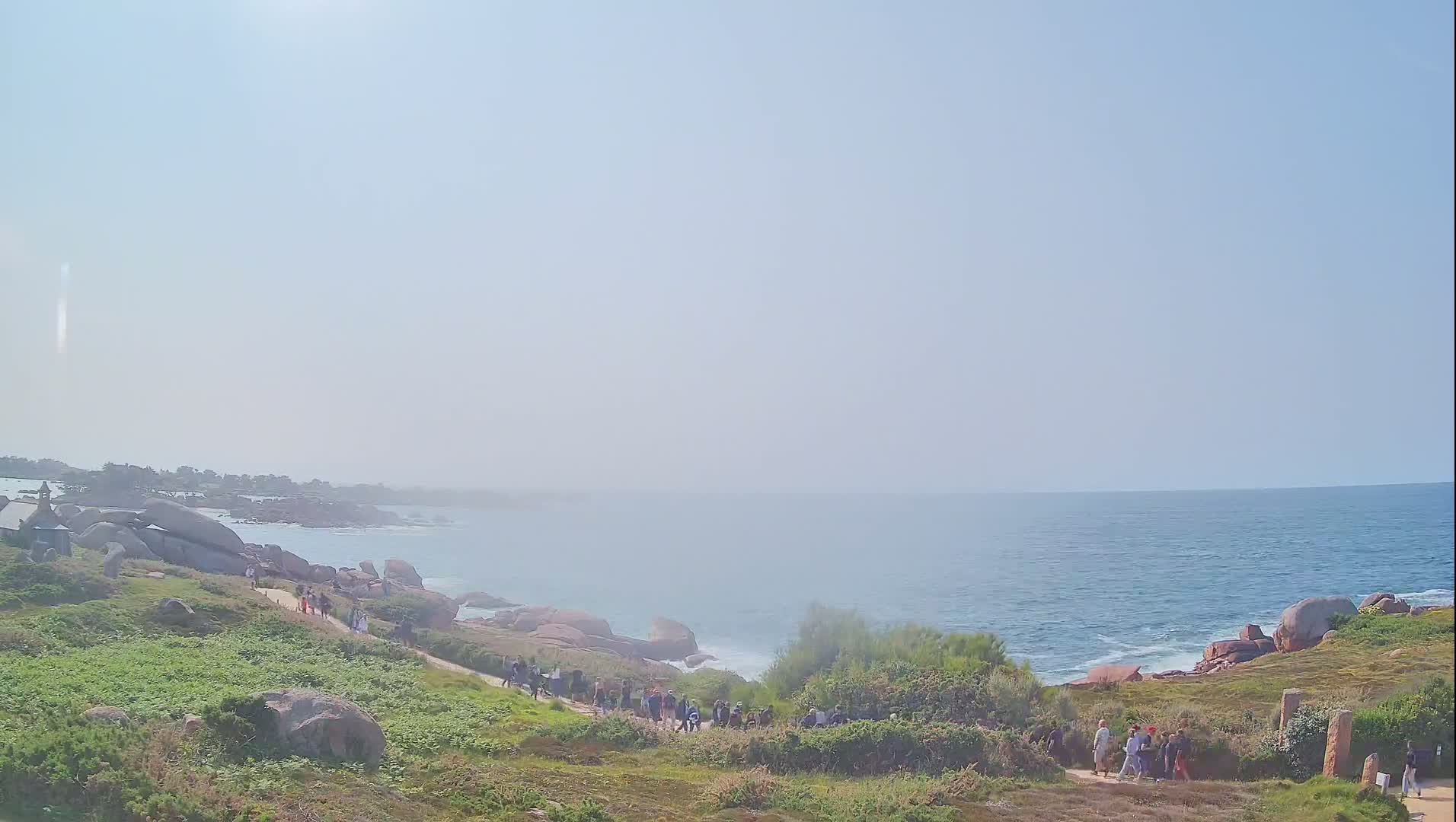 A hazy, sunny day reveals a coastal path winding through grassy terrain dotted with pink granite boulders, leading to a calm ocean with small waves.