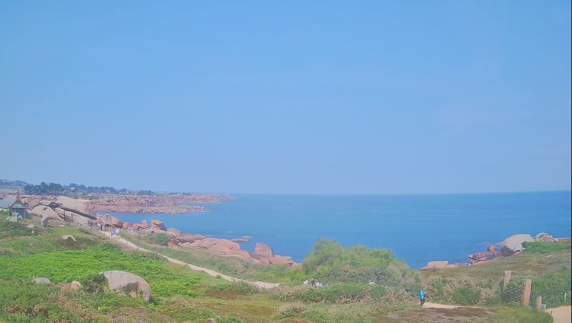 A clear, sunny day reveals a coastal scene with pink granite rocks, green vegetation, and a calm blue ocean stretching to the horizon.