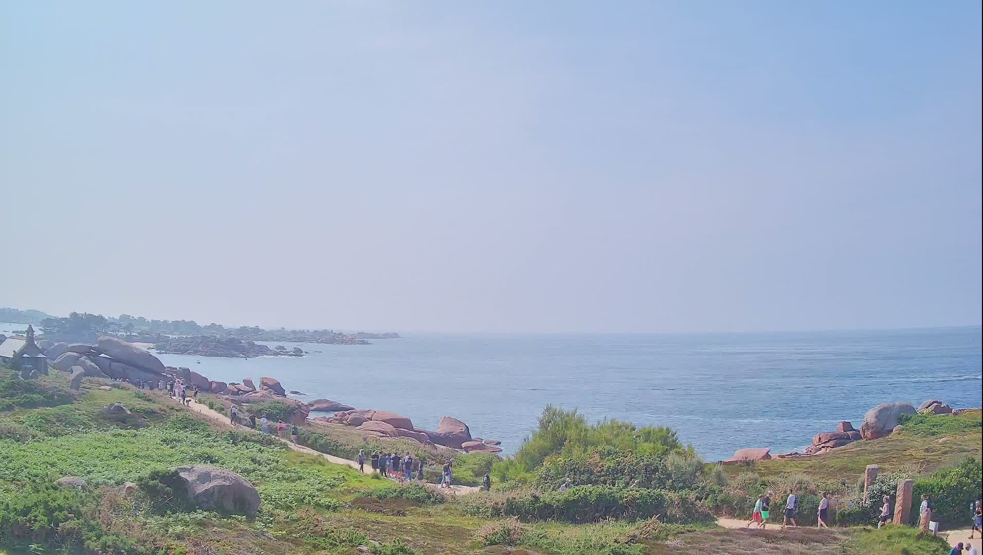 A coastal path winds through grassy terrain dotted with pink granite boulders, leading to a calm ocean under a clear, bright sky, where several people are walking.