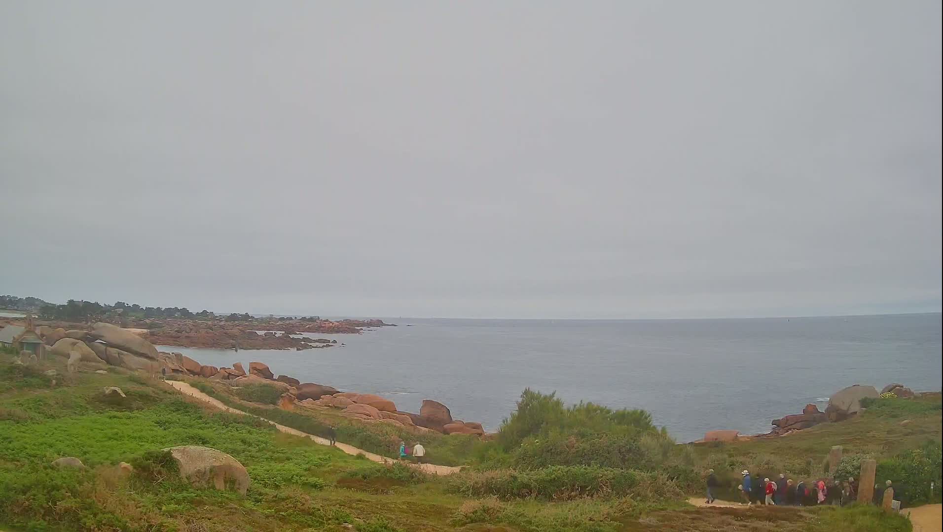 A coastal path winds through low-lying vegetation toward a calm, gray sea under an overcast sky, with pink rocks and a few people visible.
